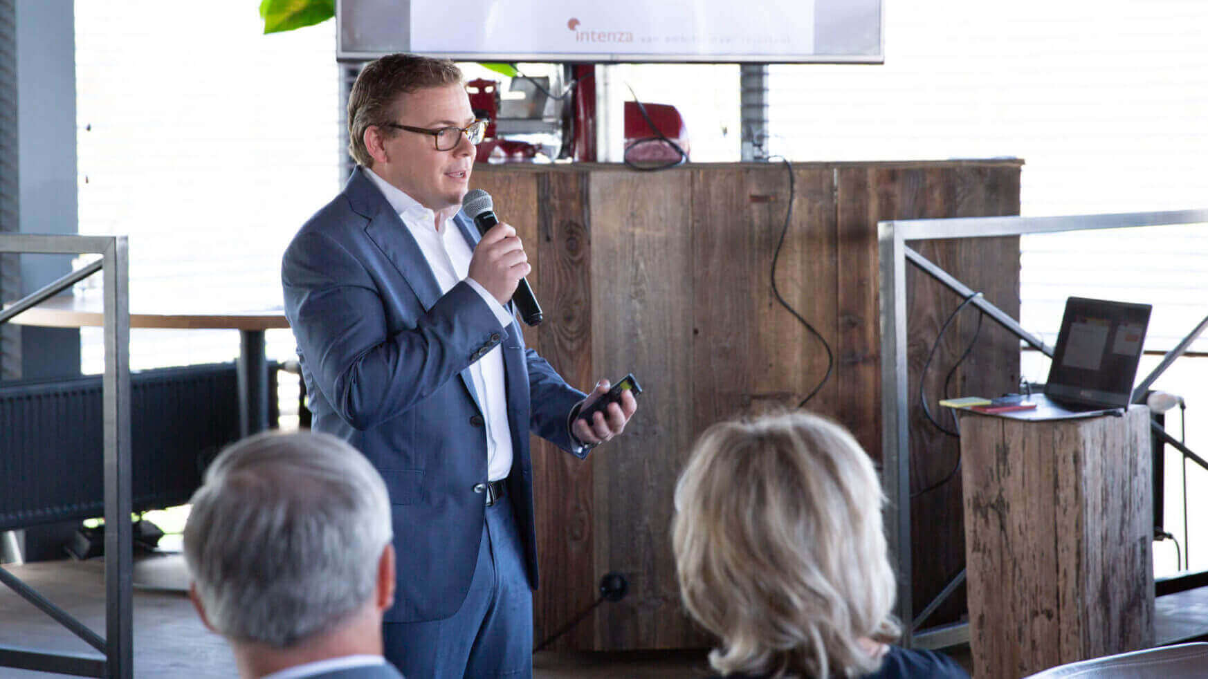 Spreker in pak tijdens een indoor evenement, spreekt het publiek toe. Het houten podiumdecor suggereert een formele setting.