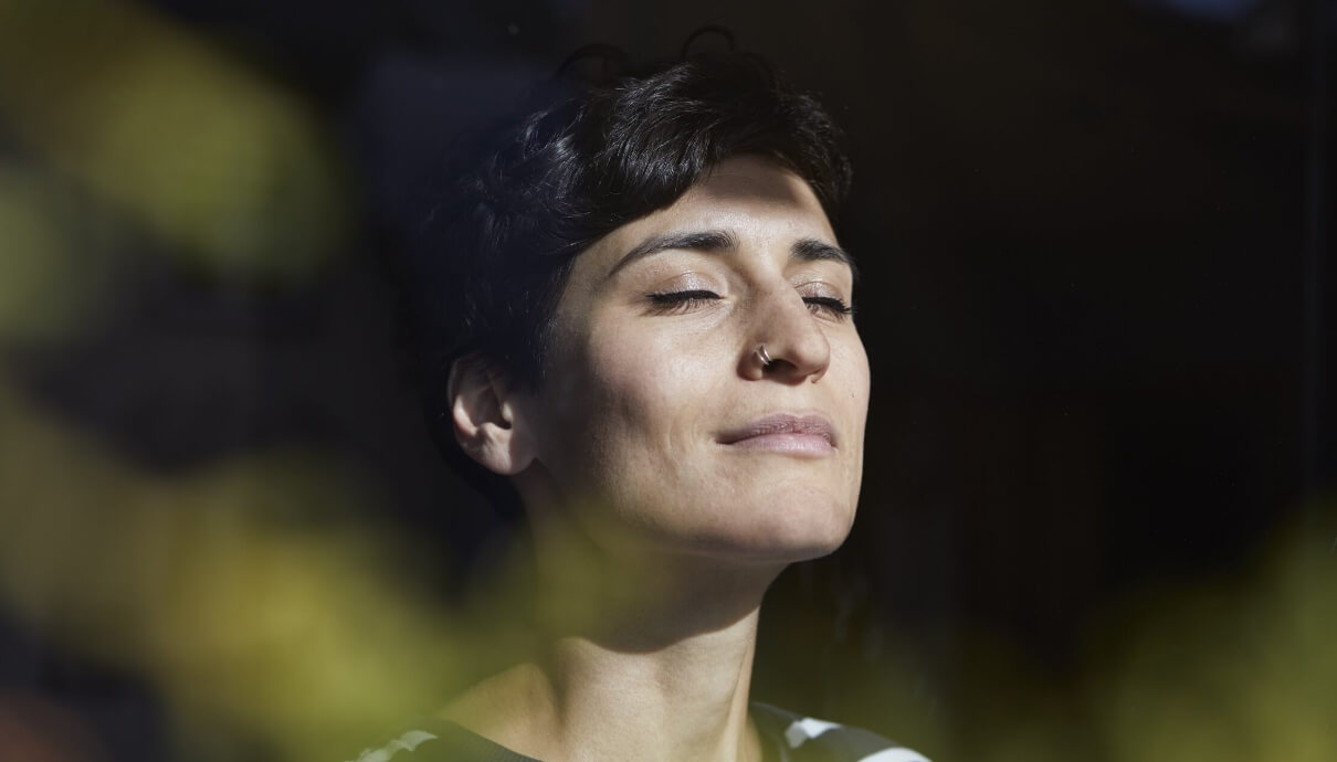 Persoon met kort haar, ogen gesloten, genietend van het zonlicht, met zachtgele bladeren wazig op de voorgrond.