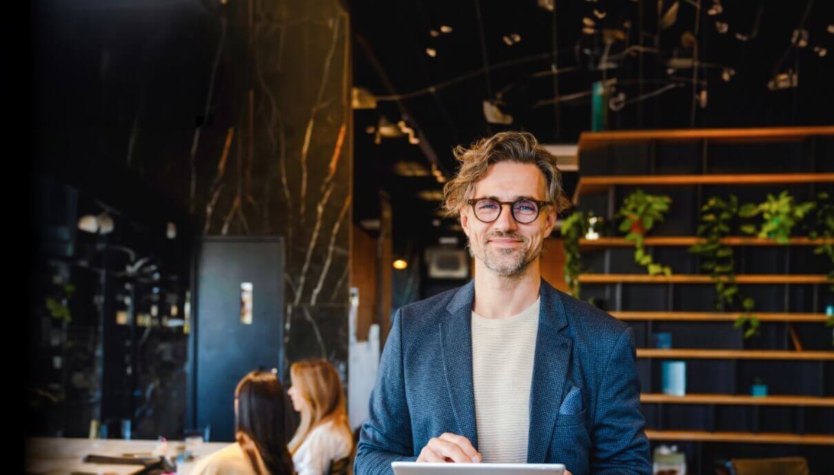 Binnen staat een lachende man met een bril en een blazer, op de achtergrond zijn mensen en planten te zien.