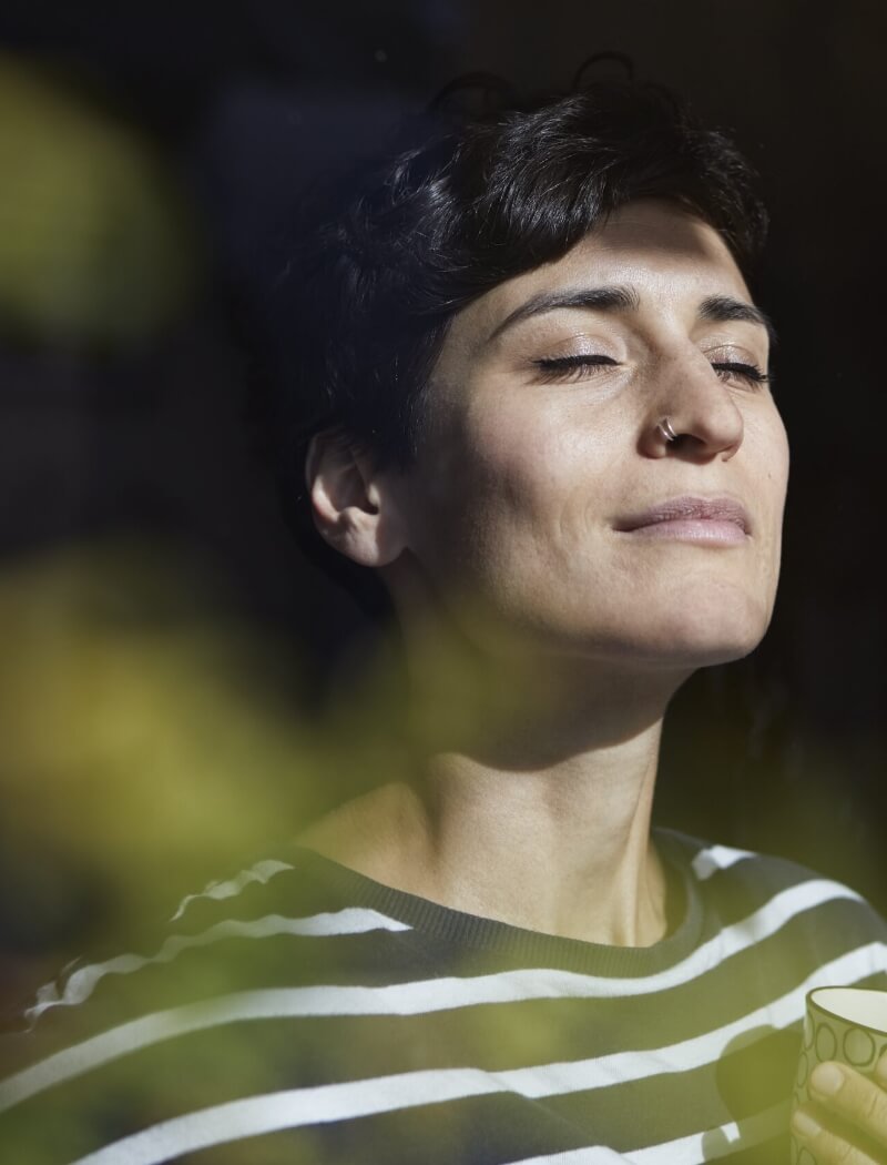 Een persoon met kort haar en strepen koestert zich in de zon, ogen gesloten, een kopje vasthoudend; vage bladeren op de voorgrond.