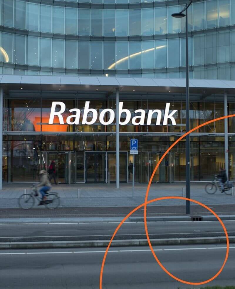 Glazen Rabobankgebouw, fietsers rijden voorbij; gebogen oranje lijnen op de voorgrond.
