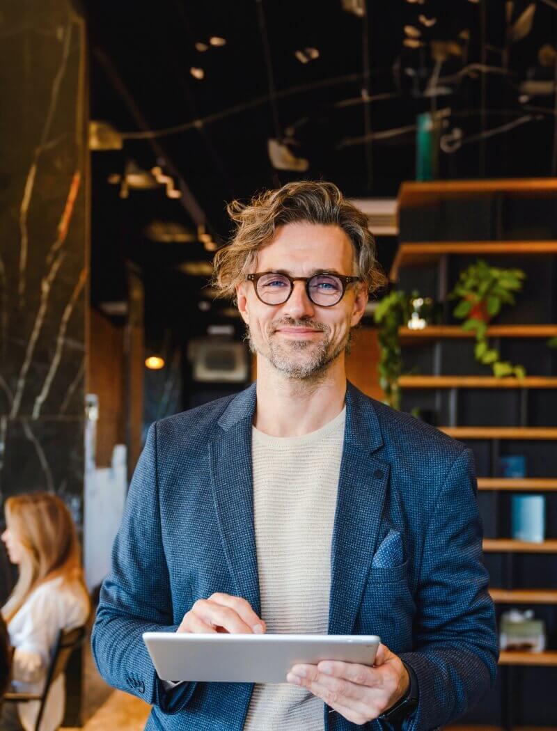 Persoon in een blauw jasje en bril, lachend met een tablet in een strak kantoor.