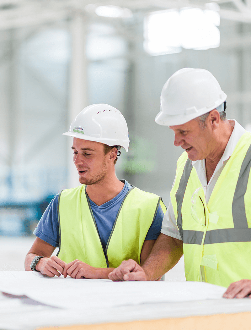 Twee medewerkers van Bouwbedrijf Vrolijk in PPE onderzoeken blauwdrukken aan een magazijntafel.