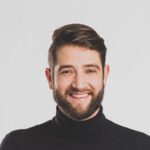 Een man met een baard in een zwarte coltrui, lachend tegen een eenvoudige achtergrond.