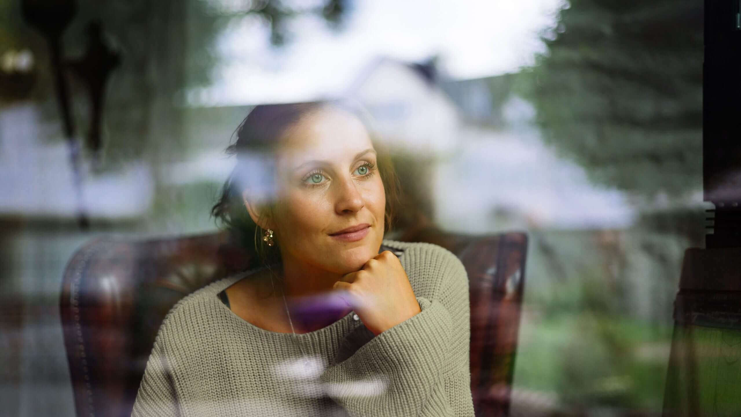 Een vrouw in een trui kijkt uit het raam, diep in gedachten verzonken, met de weerspiegelingen en het groen buiten.