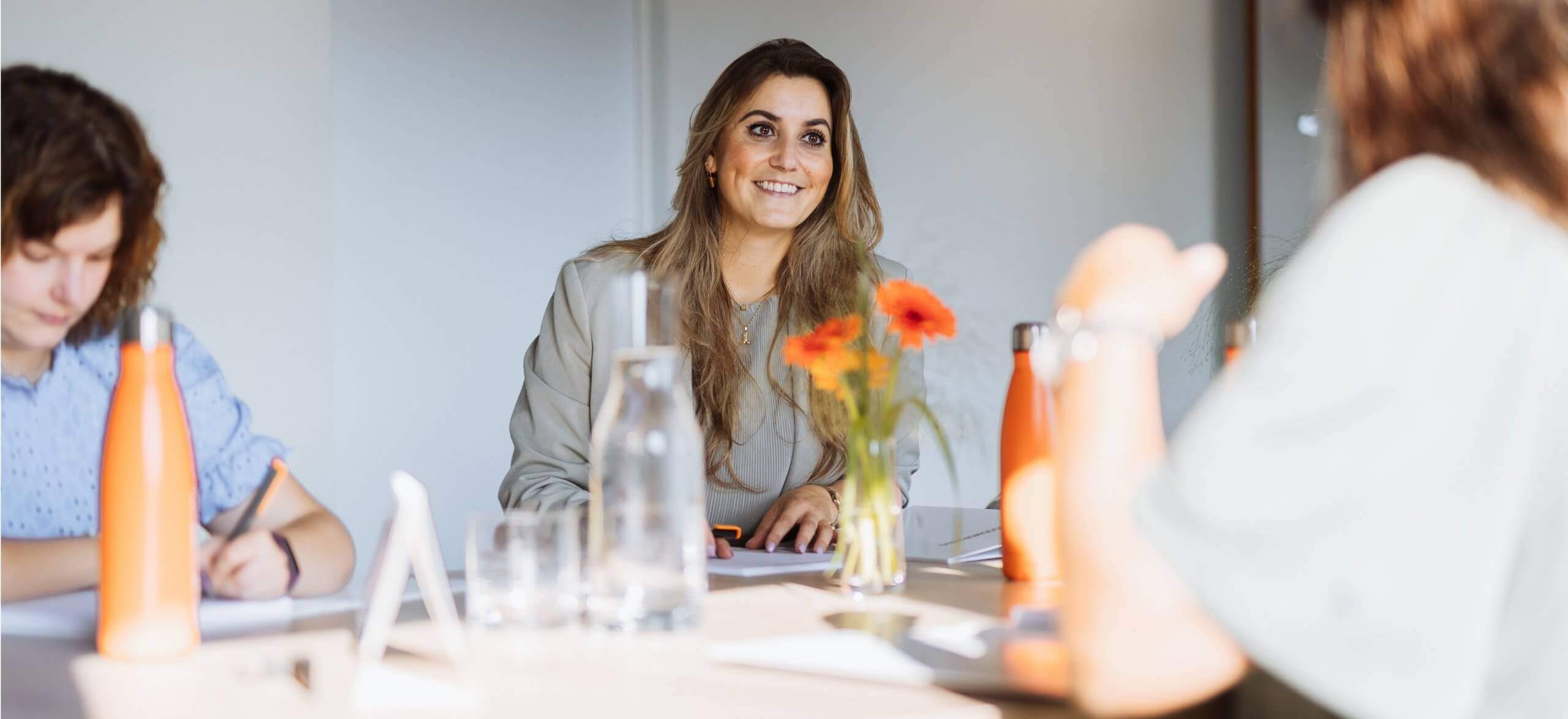 Een glimlachende vrouw zit tijdens een vergadering met papieren, een fles water en bloemen op tafel.