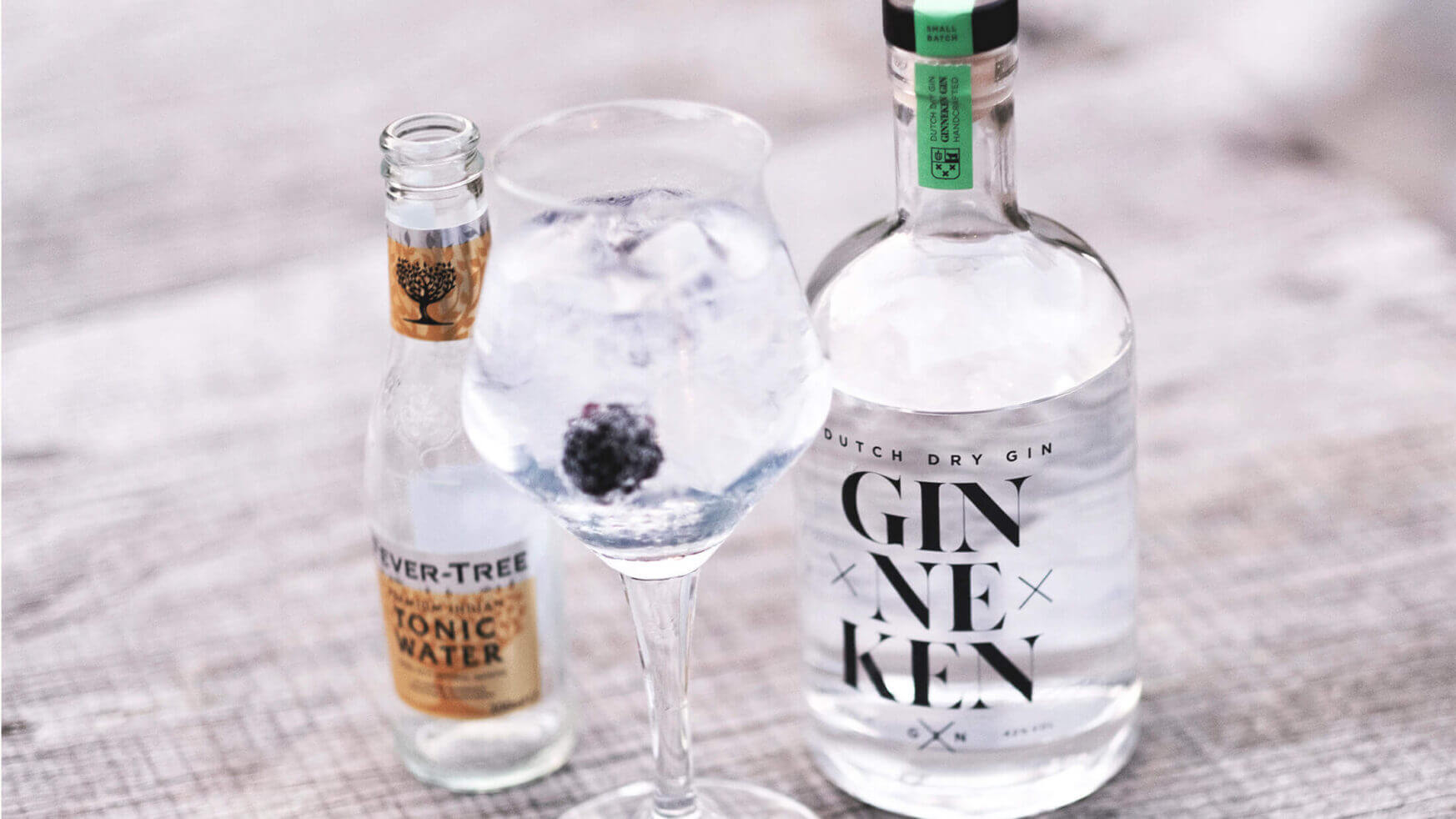 Een stijlvolle bramengin-tonic staat naast tonicwater en GinnekenGin op een rustiek houten oppervlak.