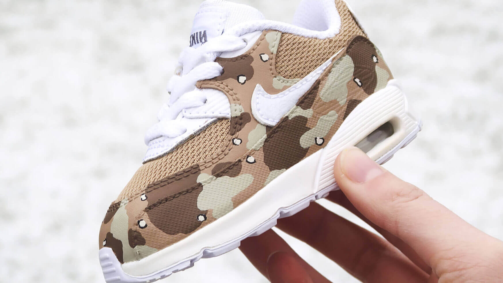 Een hand houdt een babysneaker met camouflageprint vast, met een witte swoosh en veters, speciaal gemaakt door BB.
