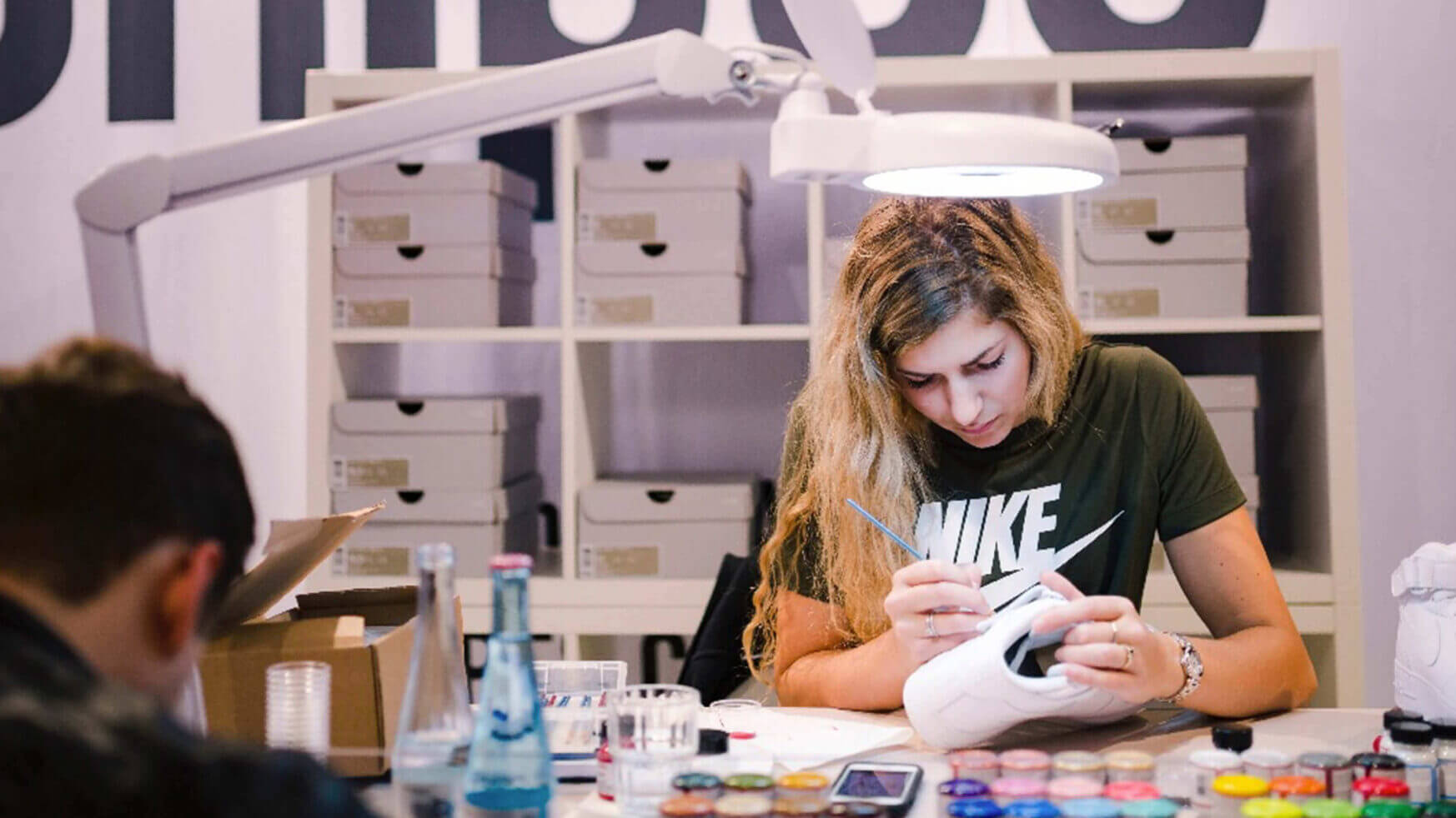 Een kunstenaar beschildert sneakers aan een felgekleurde tafel vol verf. Achterin zijn planken te zien die de chaos van "Customs by BB" laten zien.