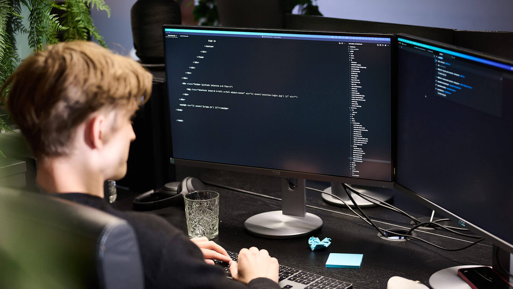 Een programmeur aan een bureau met twee monitoren, kantoorplanten en een glas water.