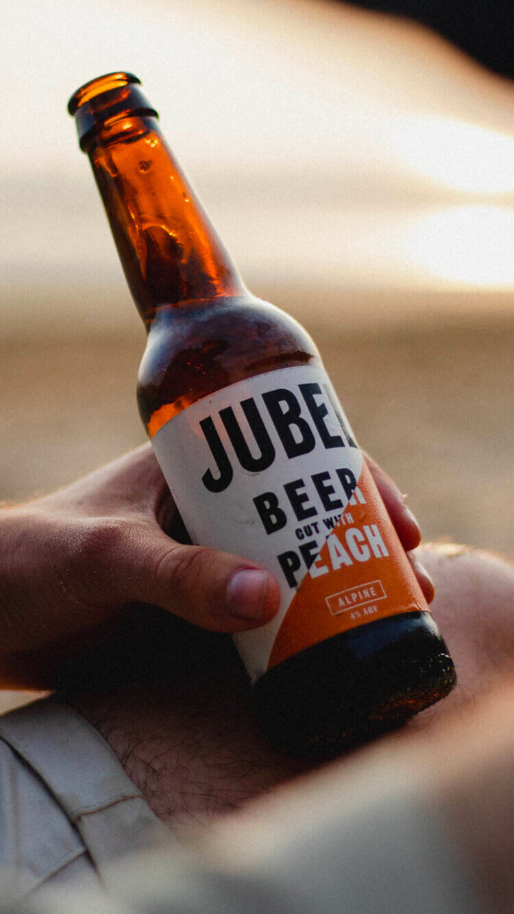 Een hand houdt een perzikkleurig Jubelbier met een oranje-wit etiket vast, dat buiten in de zon ligt te zonnen.