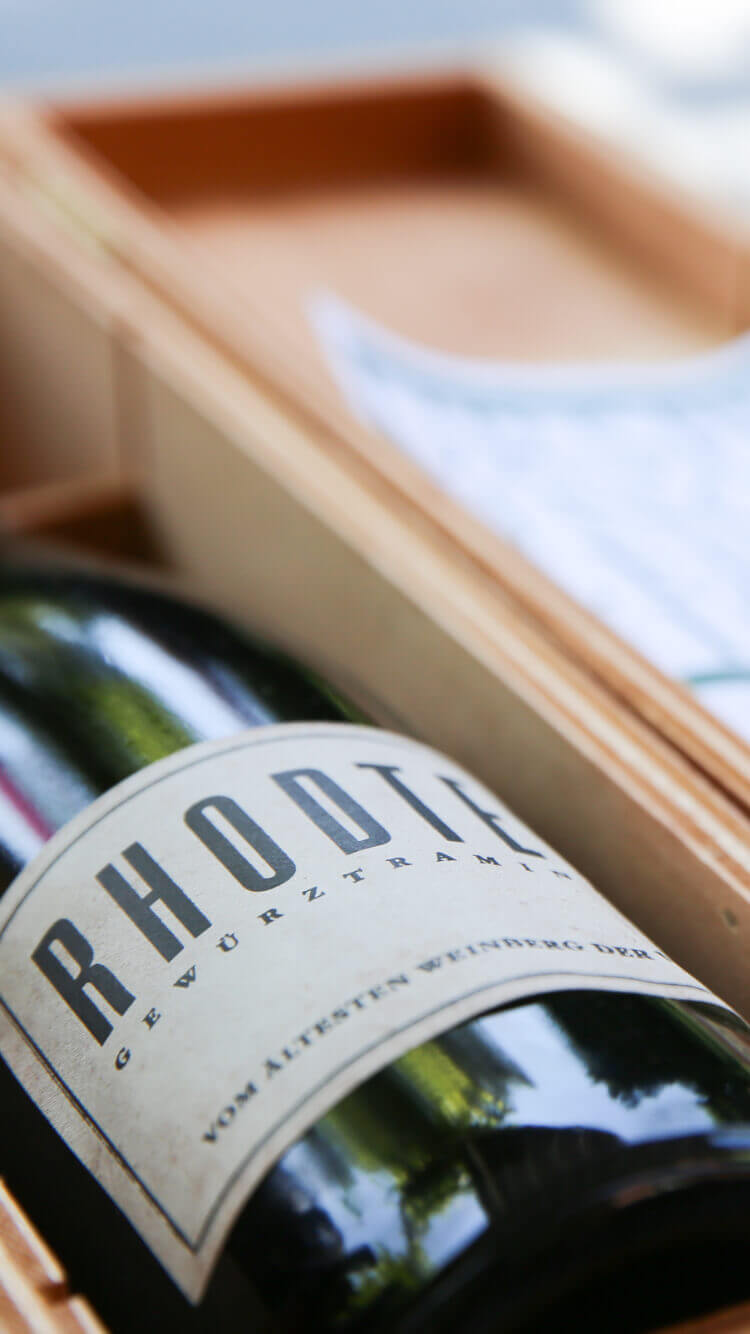 Een fles RHODT-wijn staat in een open houten kist en weerspiegelt de charme van "Les bulles enchantées" tegen een zachte, wazige achtergrond.