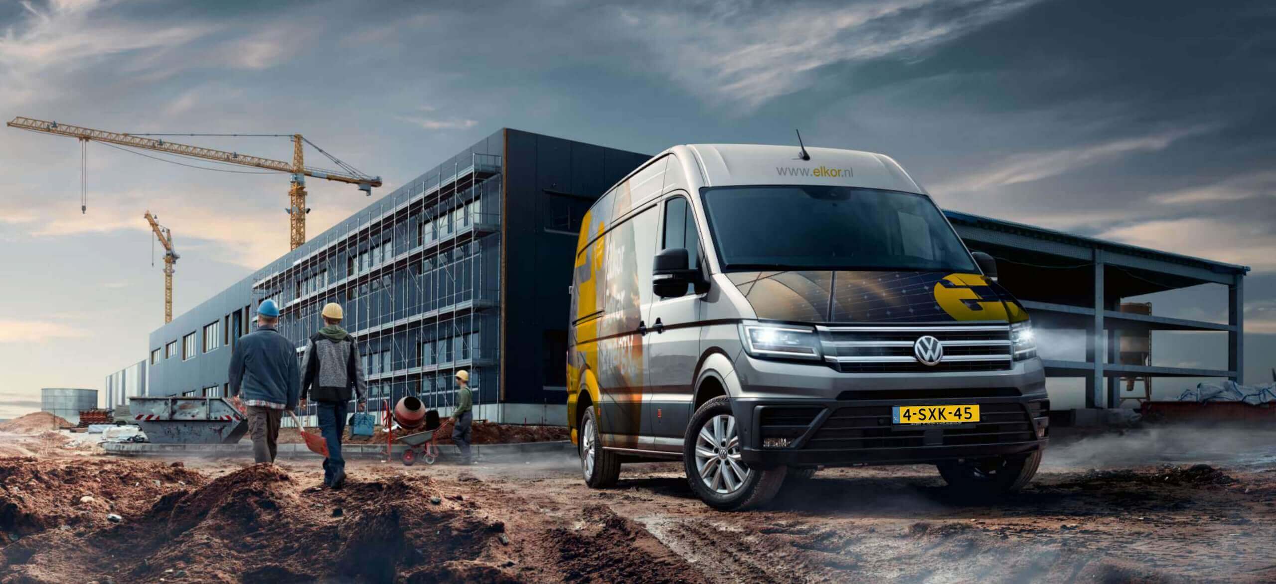 Een Volkswagenbusje met het logo van Elkor Groep staat geparkeerd op een bouwterrein met arbeiders, kranen en een gebouw.