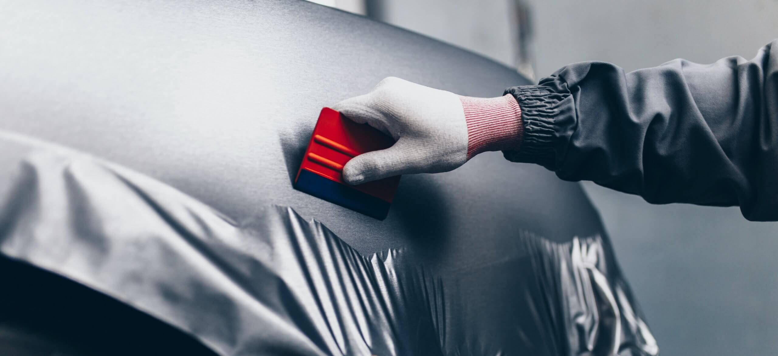 Bij Wrapgear.nl strijkt de technicus vakkundig de carwrap op metaal glad met een rood gereedschap. Hij draagt daarbij beschermende handschoenen.