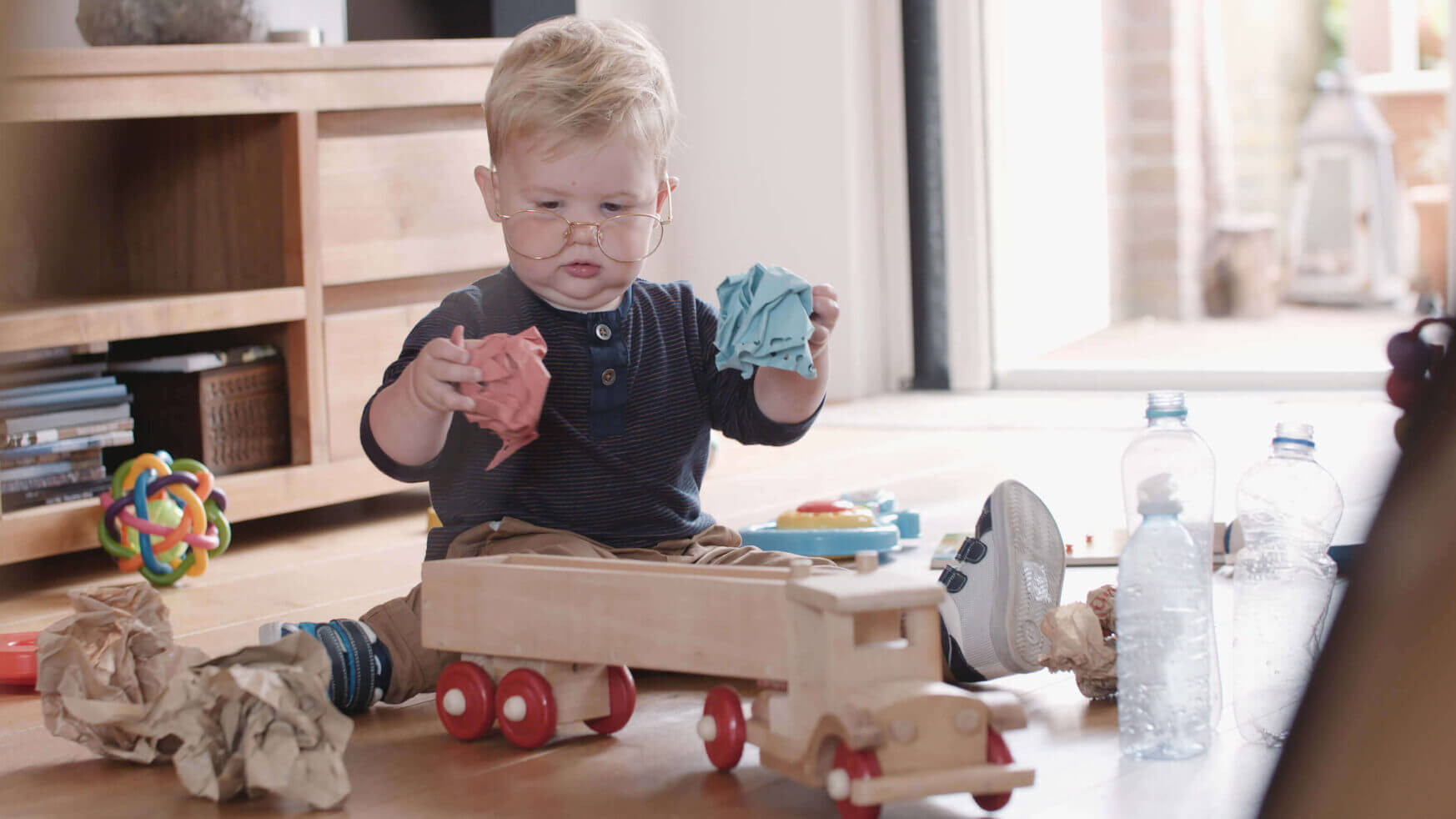 Een peuter verkent vrolijk een kamer vol speelgoed met een houten vrachtwagen en papier, wat helemaal in de sfeer van "Samen Aan De Bak!" komt.