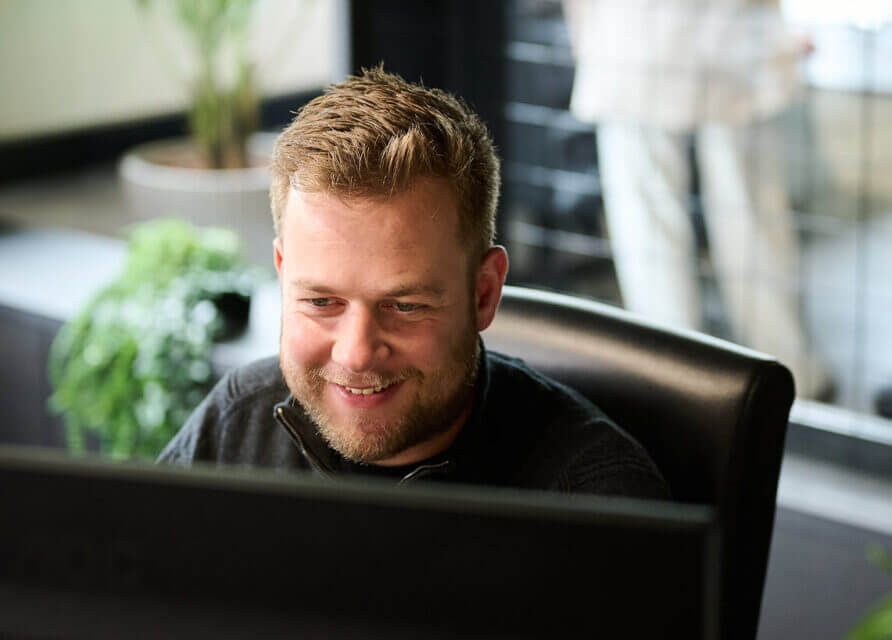 Een man grijnst naar zijn computer in een kantoor vol planten. Hij straalt succes uit.
