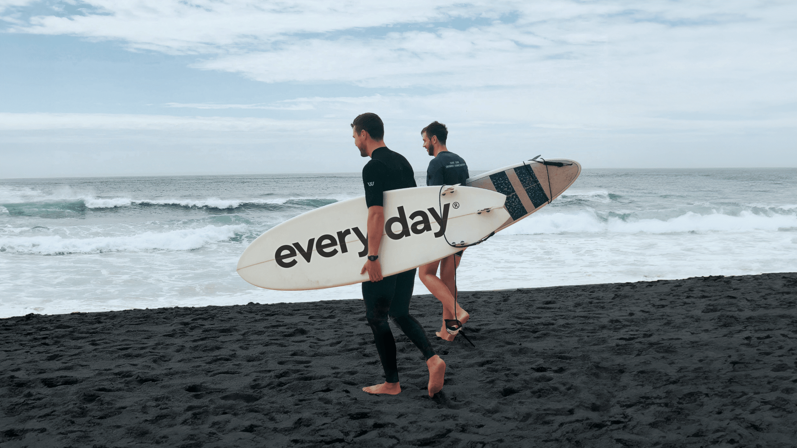 Twee surfers wandelen over een zwart zandstrand, met een surfplank in hun handen met het opschrift 'elke dag'.