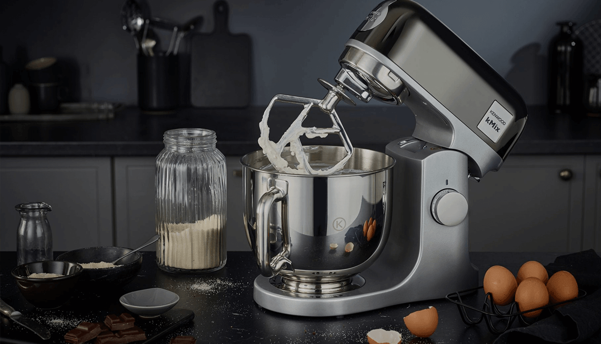 Een standmixer met slagroom, eieren en bloem staat op een donker aanrecht.