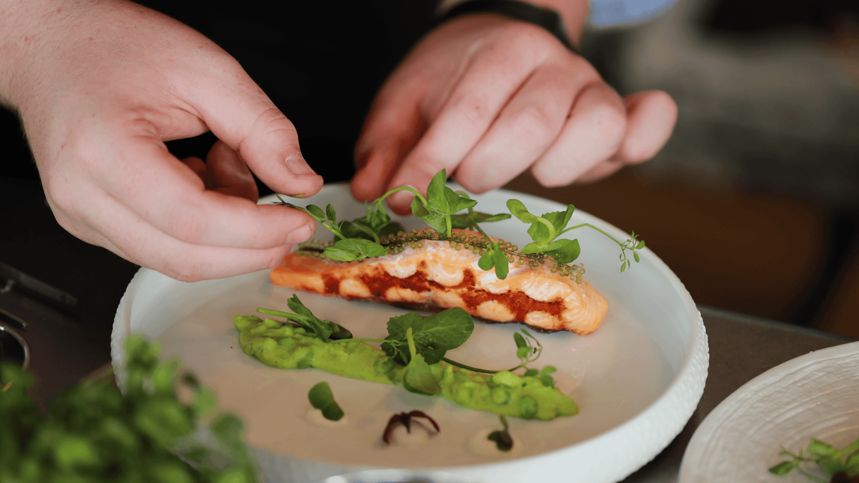 Hands topping zalmfilet met verse groenten en tuinkers, geserveerd met erwtenpuree op een wit bord.