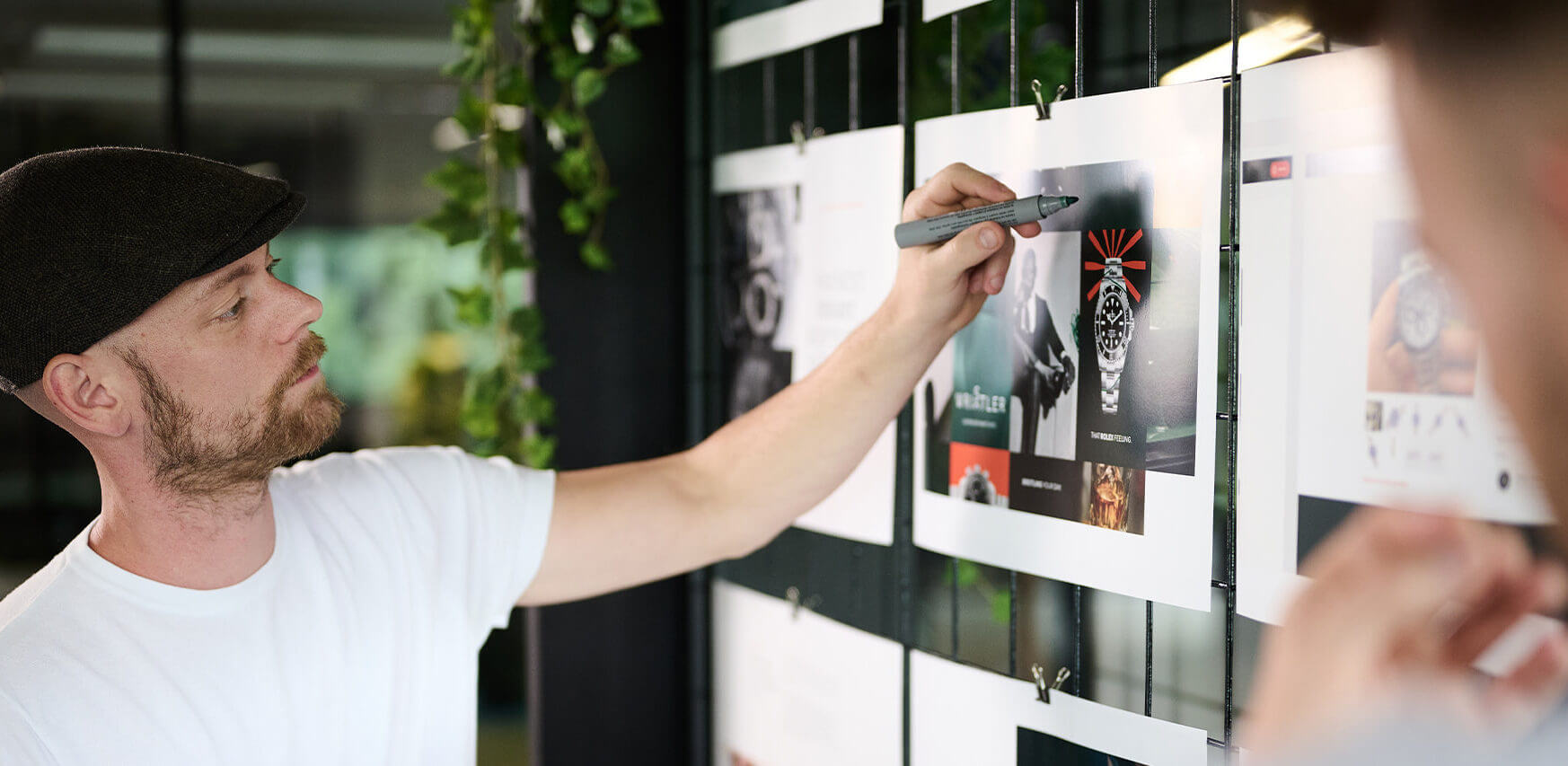 Een man met een pet schetst druk ideeën op een ontwerp dat op de rastermuur is geprikt.