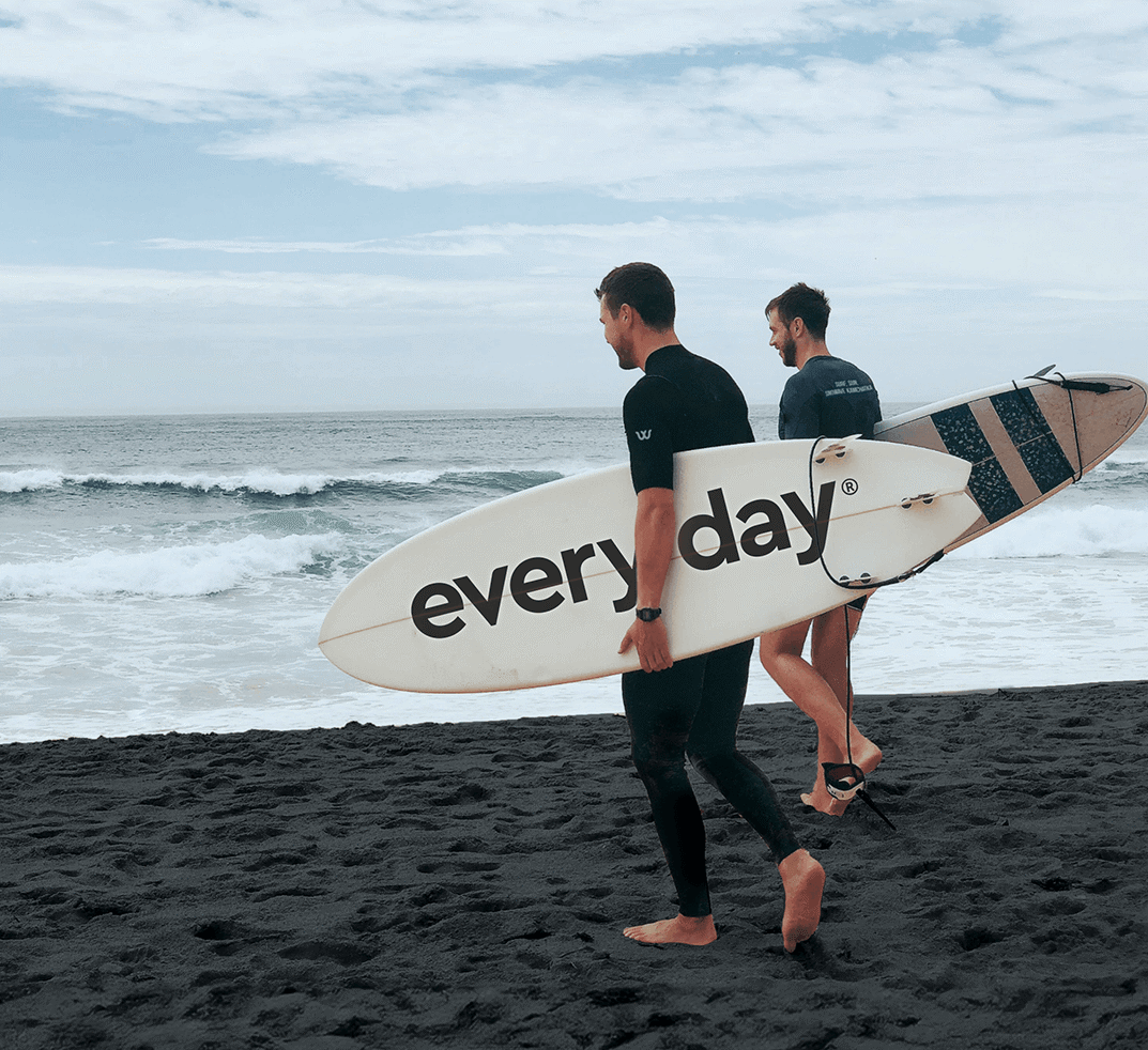 Twee surfers gaan naar de oceaan. De één houdt een bord vast met het opschrift 'elke dag'.