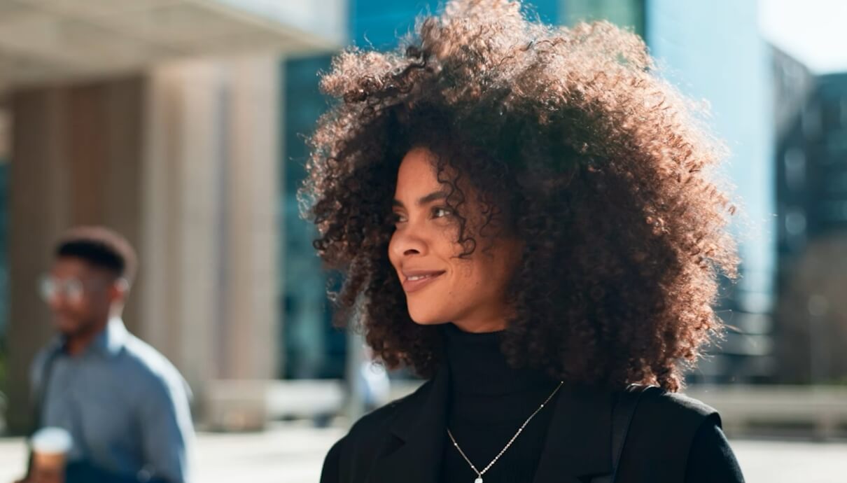 Zelfverzekerde, krullende vrouw in een zwarte coltrui lacht te midden van de stad. Ze vertegenwoordigt met trots FIT Group.