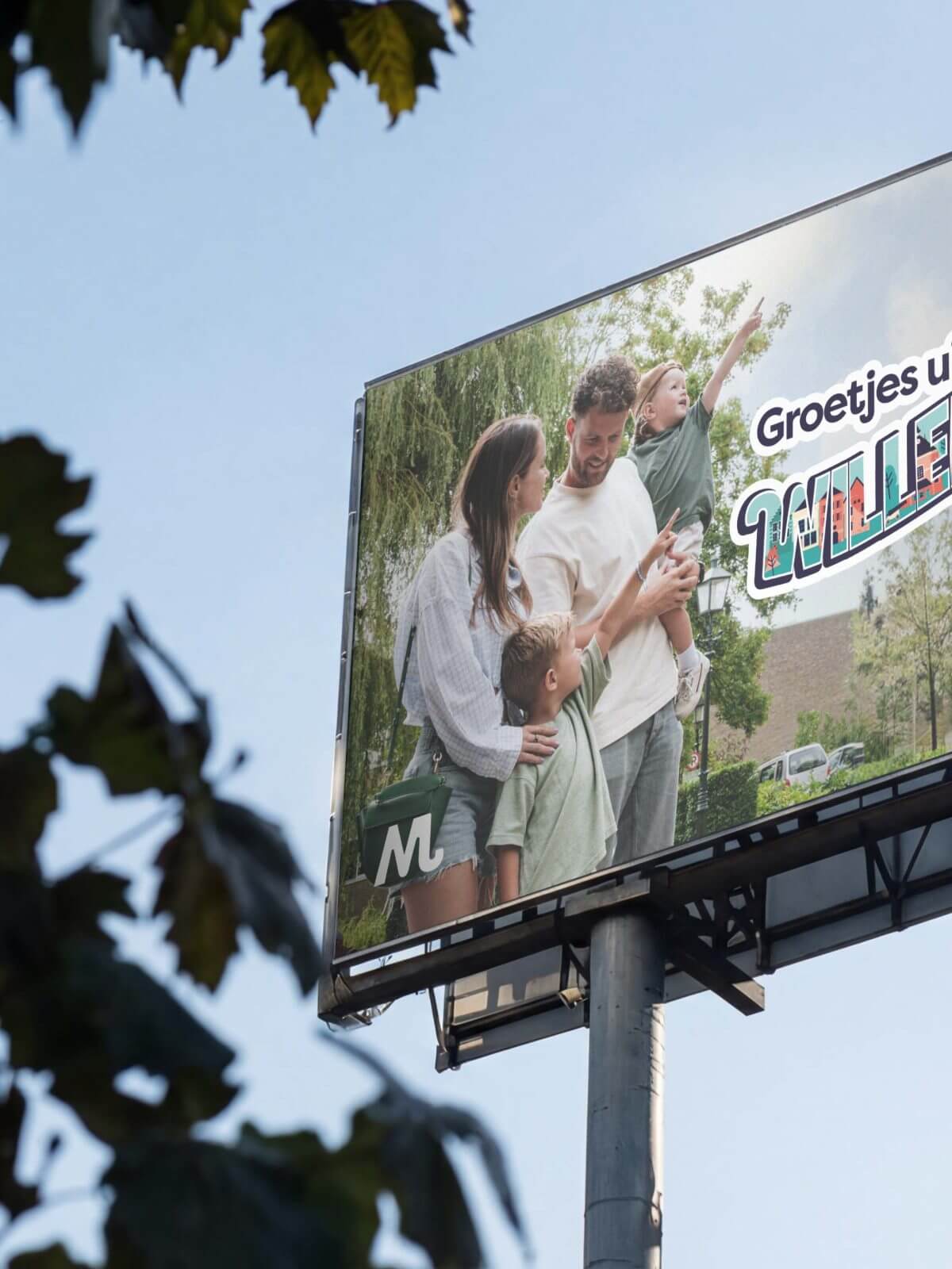 Billboard met een gelukkig gezin van vier, blauwe lucht en tekst: "Groetjes uit HEMAWIJKSTAD. Bezoek Moerdijk!.
