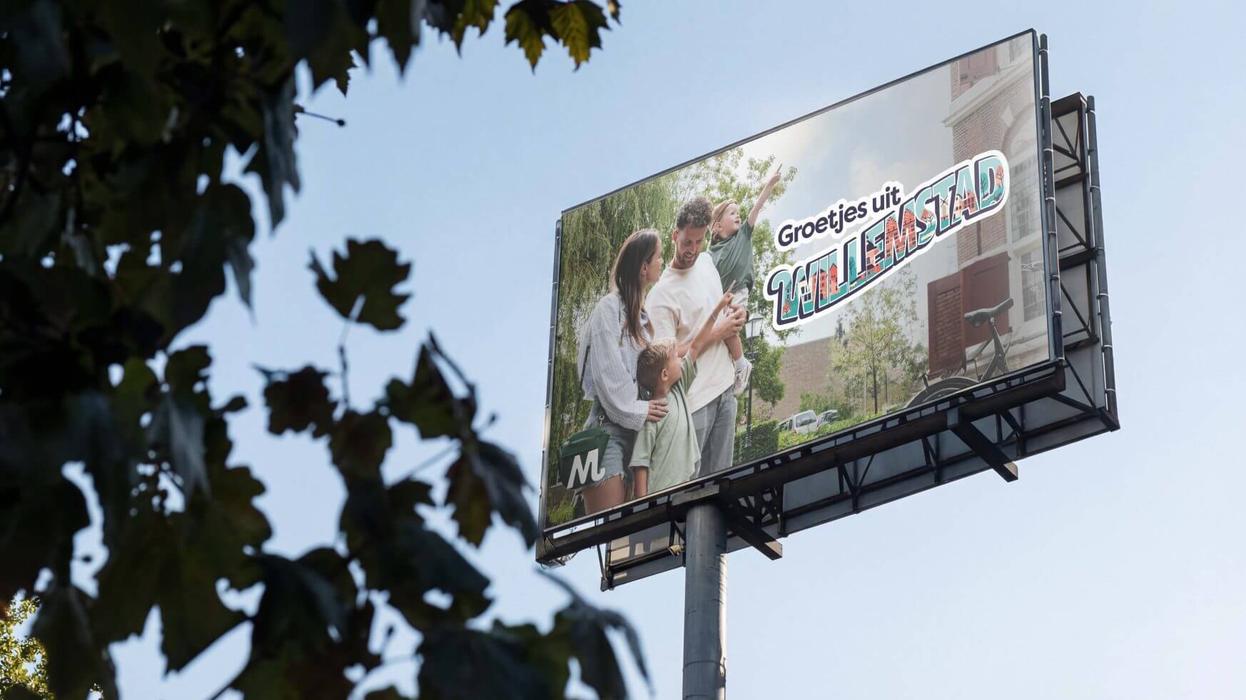 Billboard met een gelukkig gezin van vier, blauwe lucht en tekst: "Groetjes uit HEMAWIJKSTAD. Bezoek Moerdijk!.