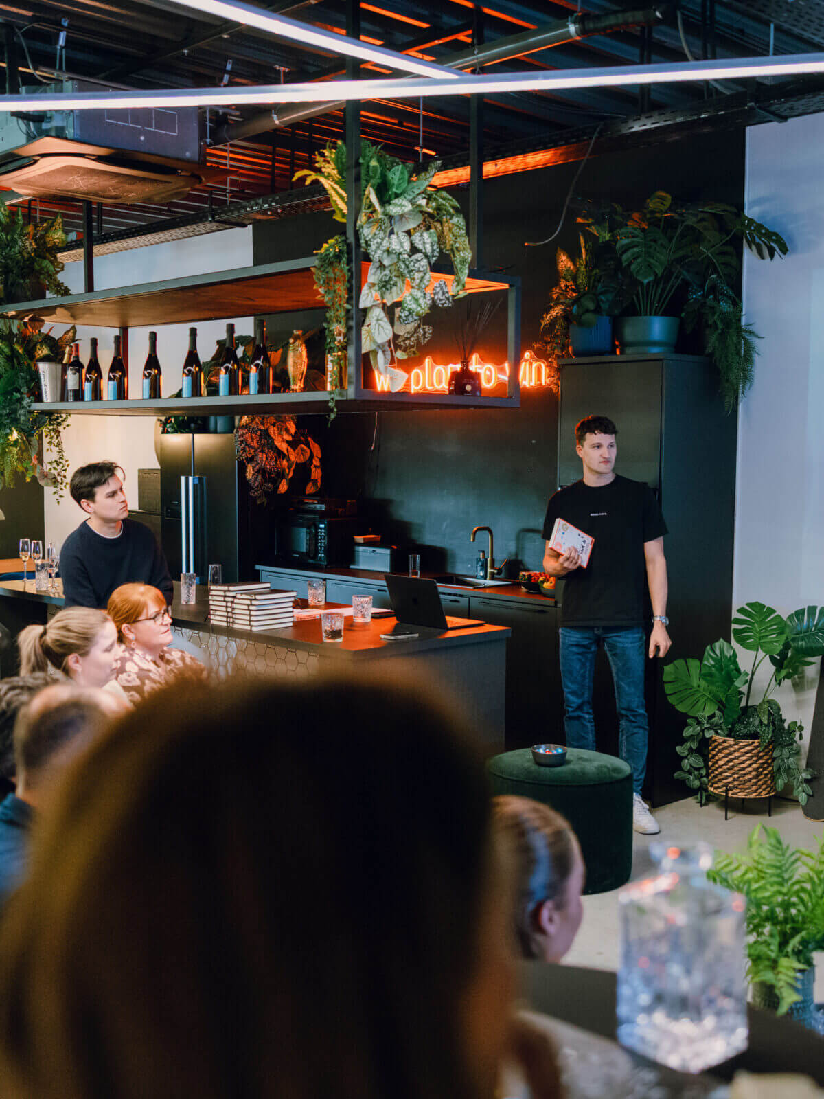Een man presenteert aan een zittende groep in een stijlvolle, met planten gevulde ruimte met een groot scherm.