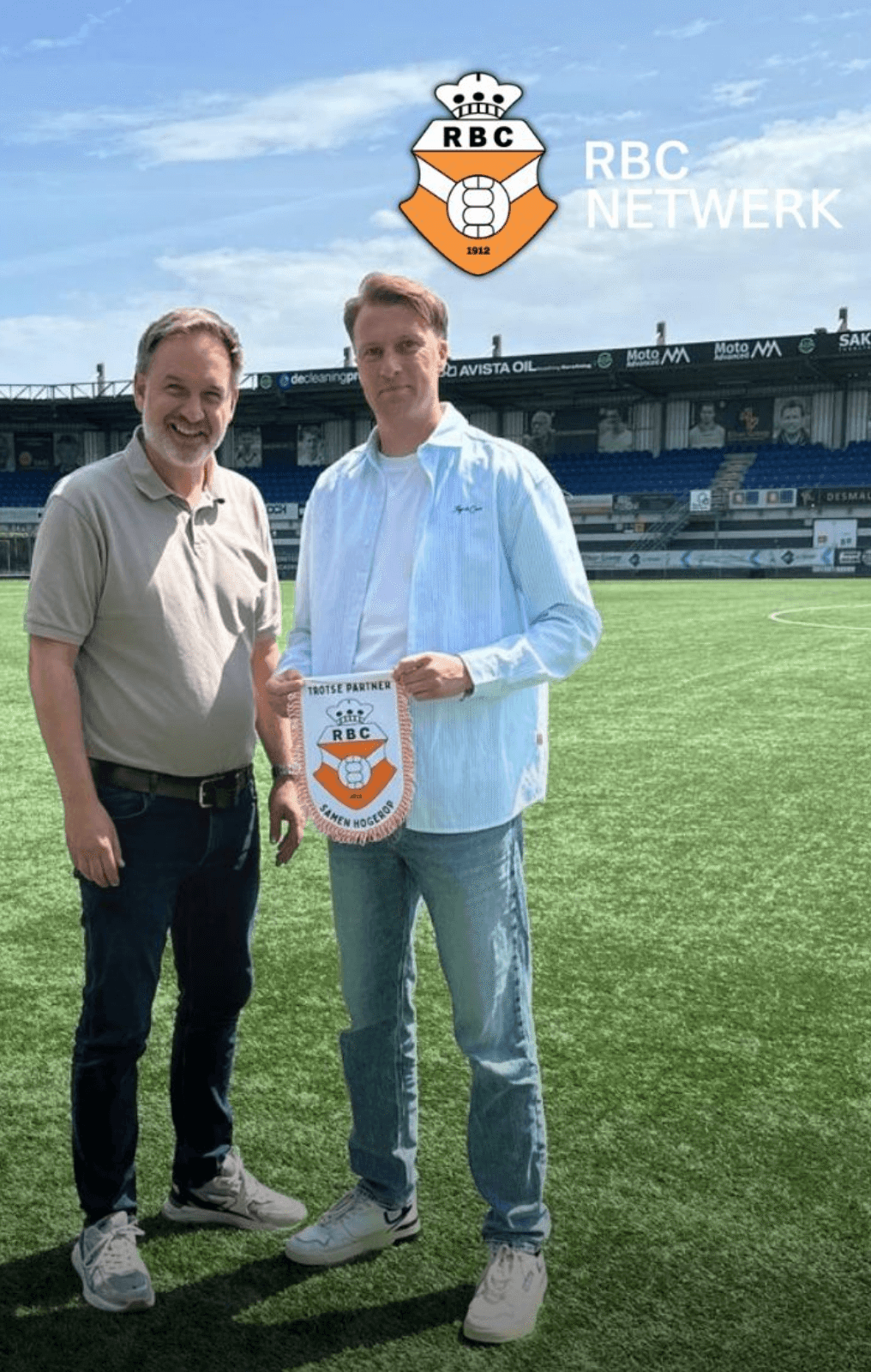 Twee mannen op een voetbalveld houden een RBC Netwerk bord vast, stadion en logo zichtbaar op de achtergrond.