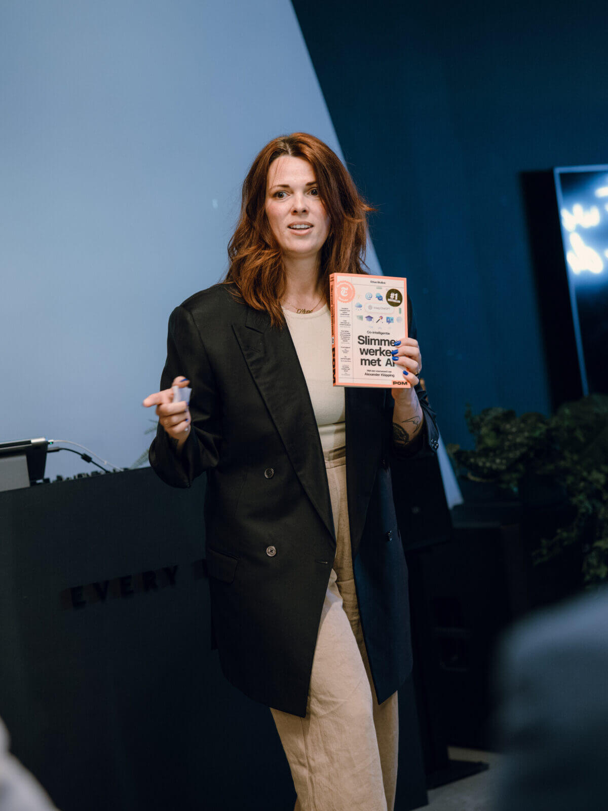 Vrouw presenteert aan een publiek, houdt een boek vast, met een presentatiescherm op de achtergrond.
