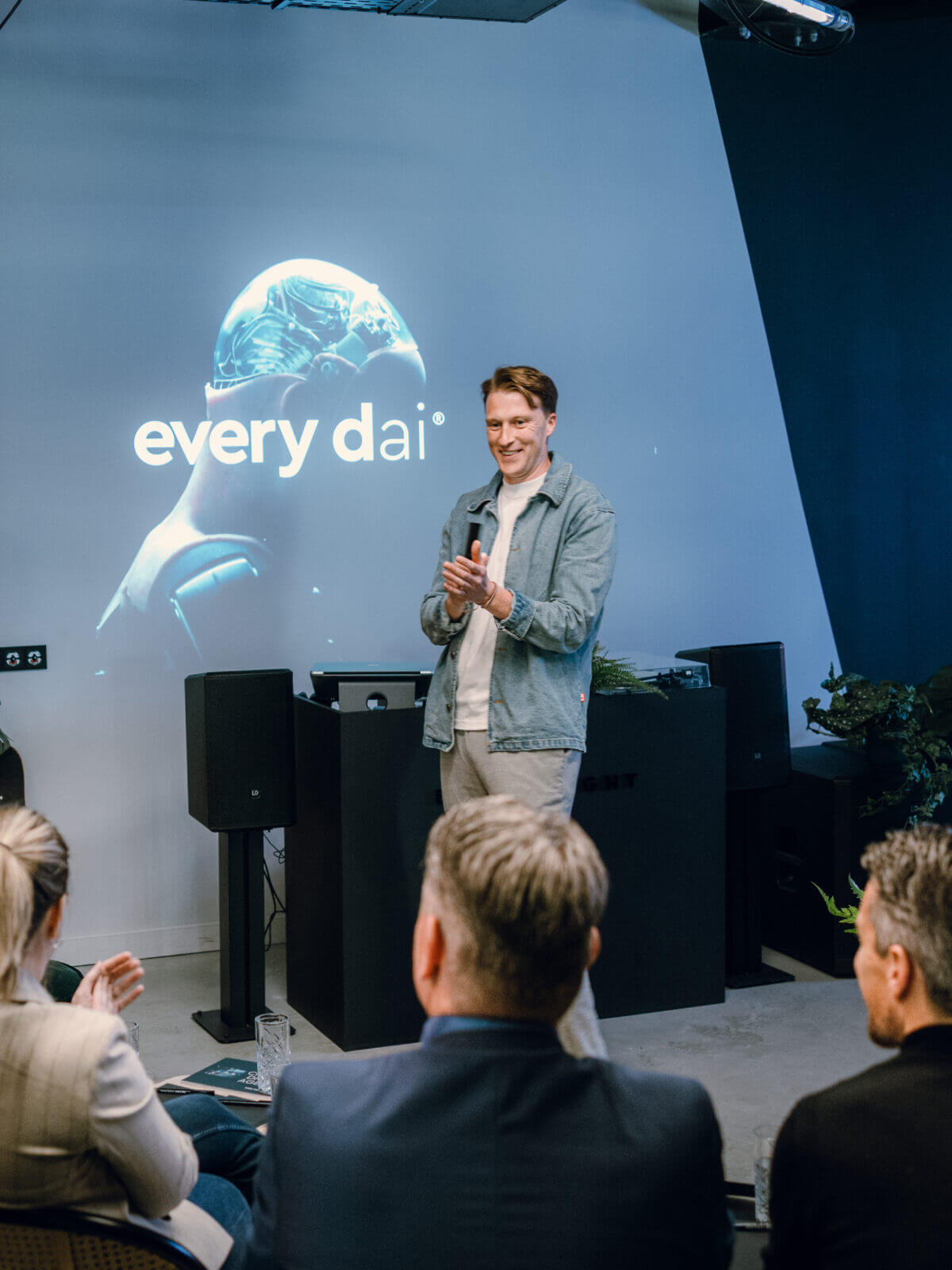 Man spreekt een publiek toe in een strakke kamer, met "every dai" achter hem geprojecteerd.