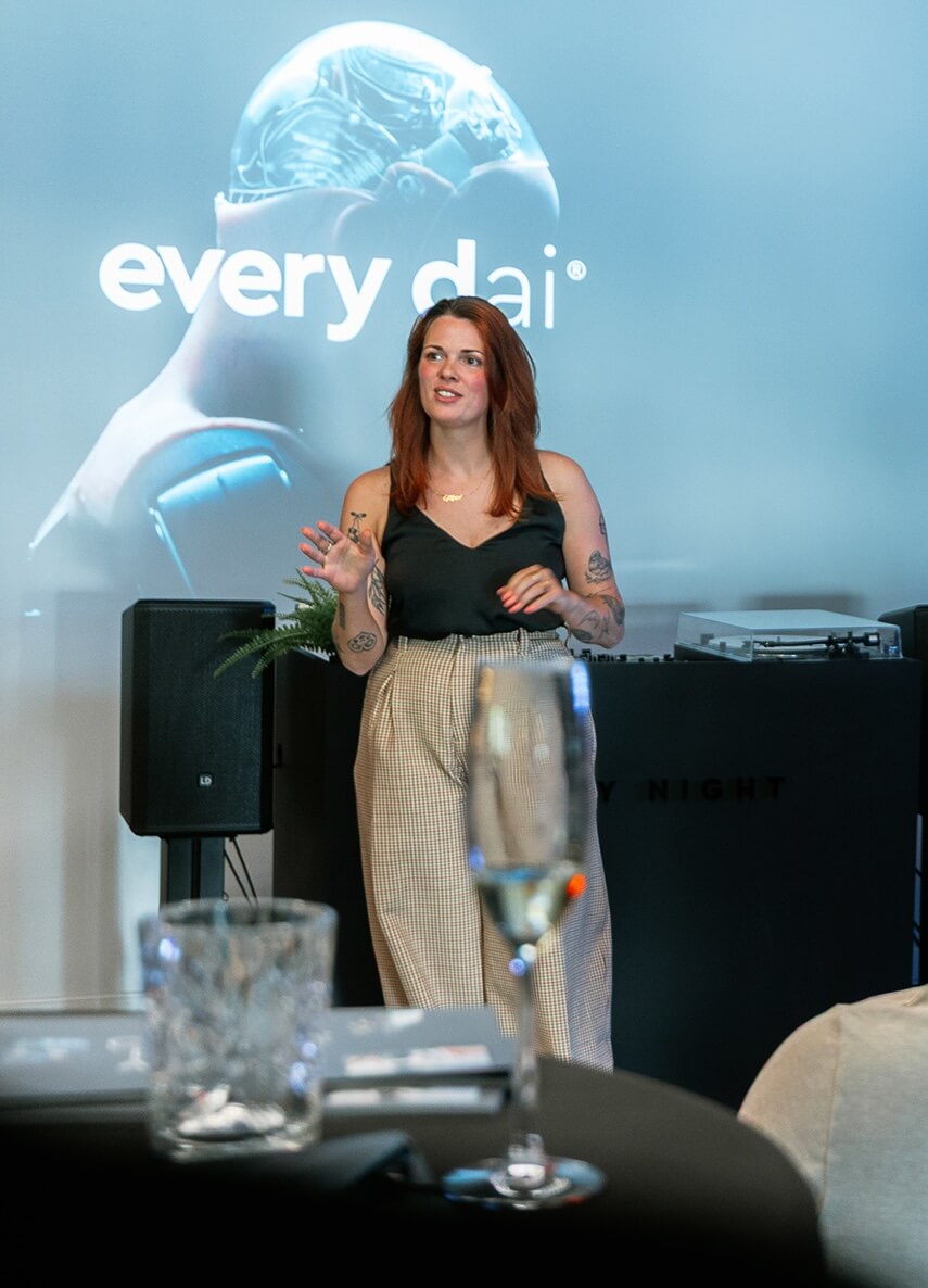 Vrouw spreekt op het podium; achter haar toont een scherm "every dai" en een robotbeeld.