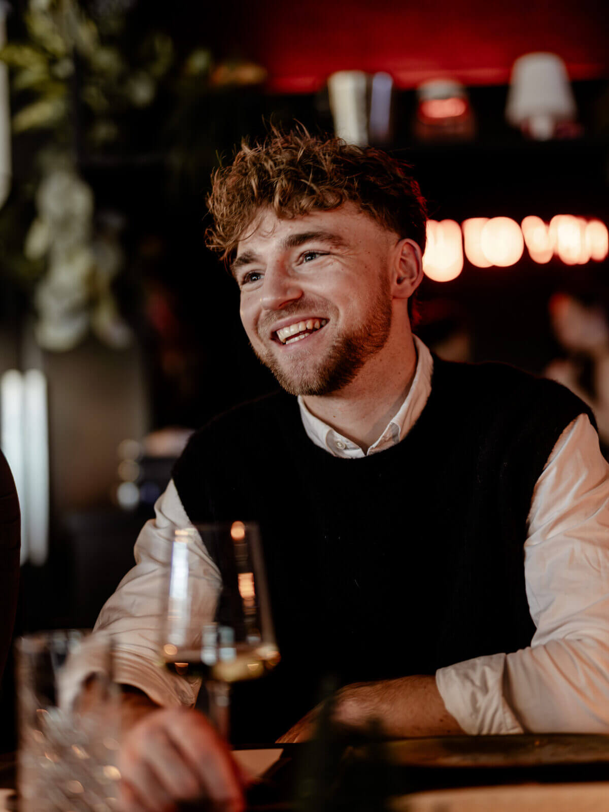 Een man met krullend haar glimlacht aan een tafel in een donker verlicht restaurant.