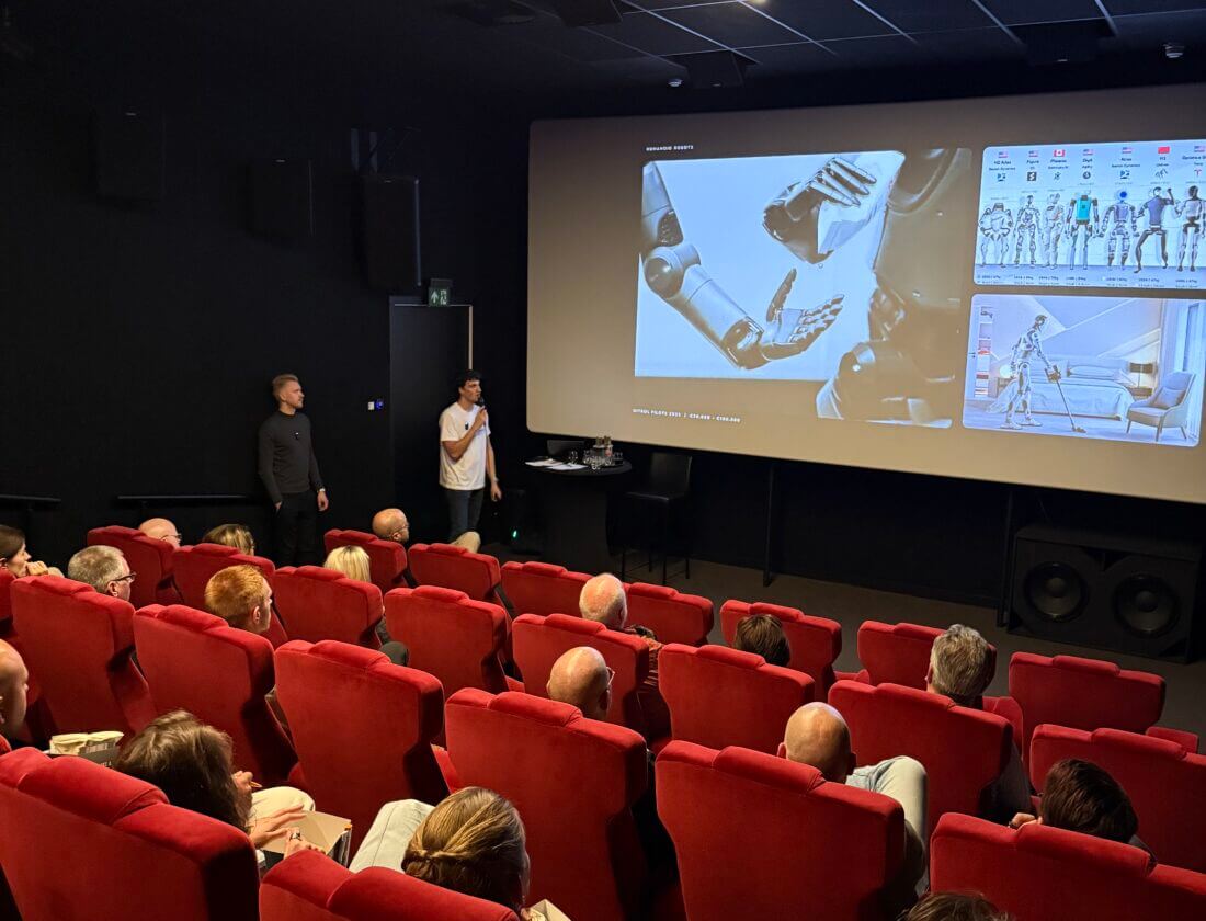 Theaterpubliek kijkt naar een techniek- en robotpresentatie op een groot scherm vooraan.