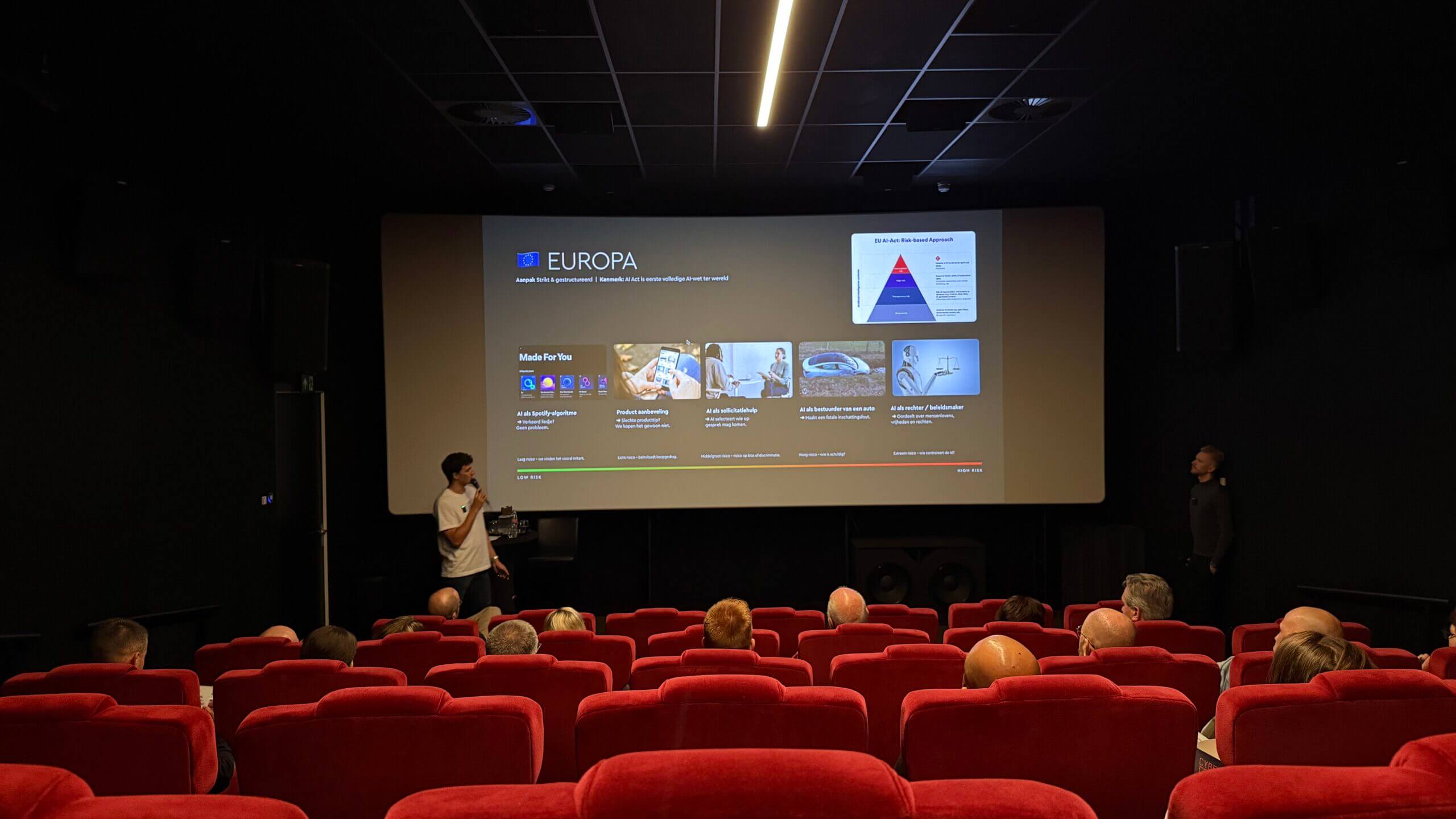 Het publiek bekijkt een presentatie op een groot scherm in een donker theater met rode stoelen.