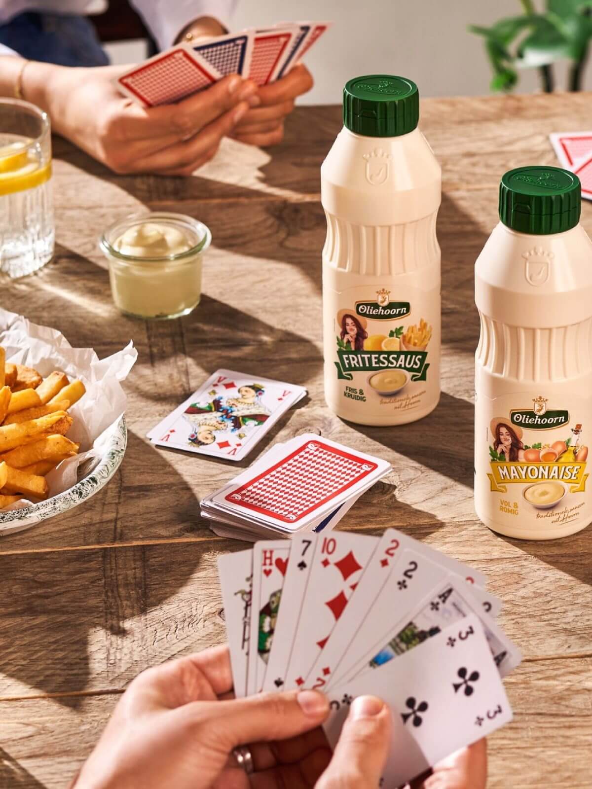 Vier vrienden spelen kaart aan een houten tafel bedekt met friet, sausjes, drankjes en gefrituurde snacks.