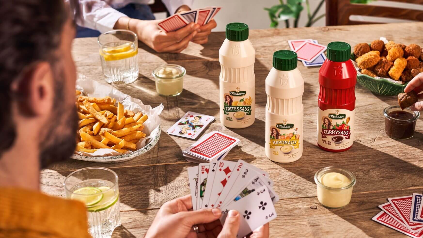 Vier vrienden spelen kaart aan een houten tafel bedekt met friet, sausjes, drankjes en gefrituurde snacks.