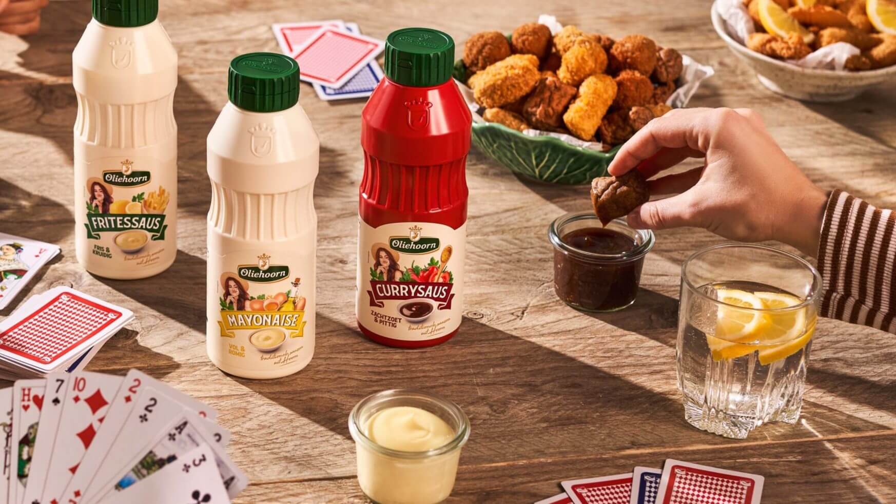 Sauzen, snacks en speelkaarten op een houten tafel, handen reiken naar eten en kaarten.