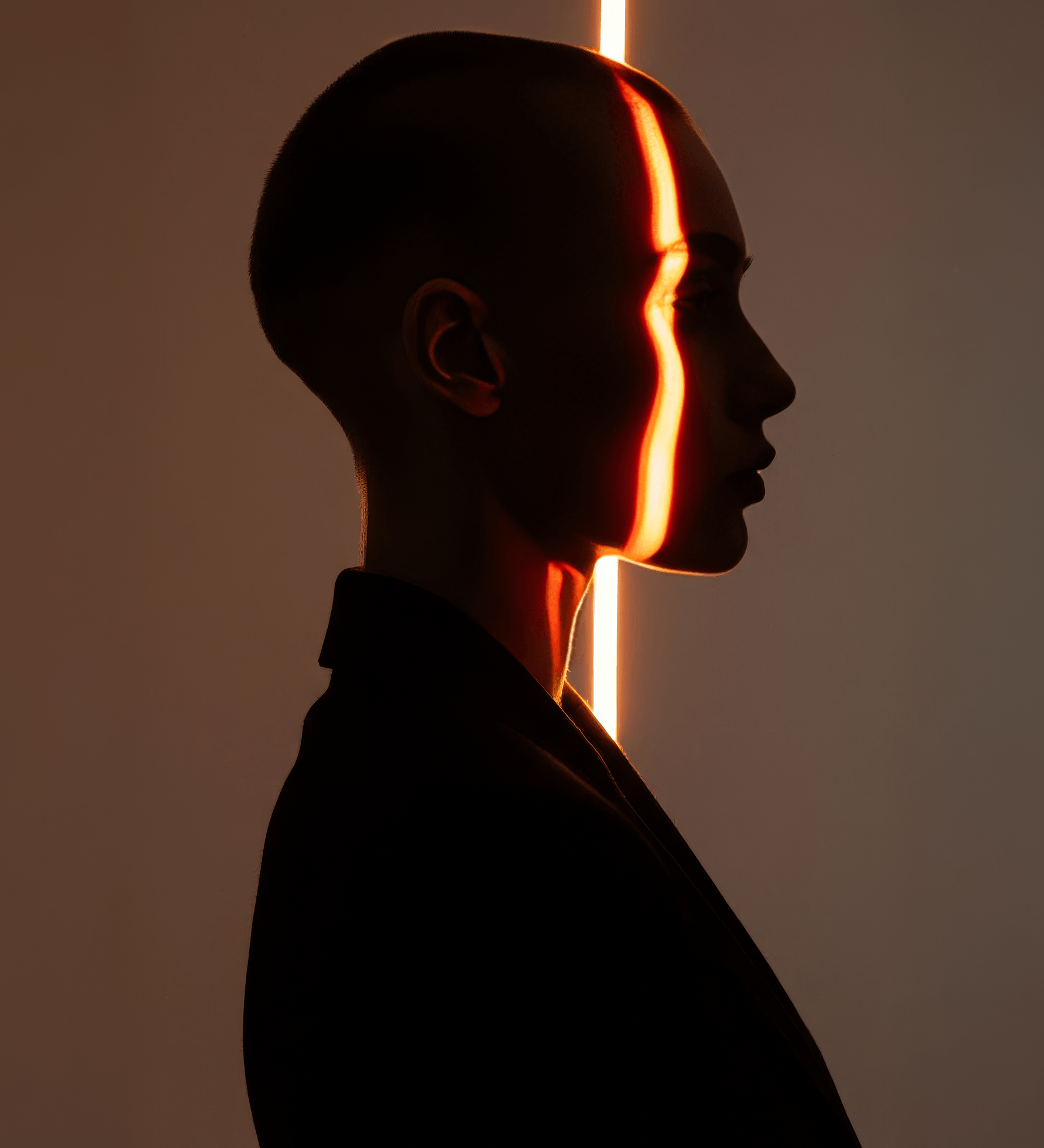 Profiel met geschoren hoofd, dagelijks verlicht door een verticale strook warm licht op het gezicht, tegen een donkere achtergrond.