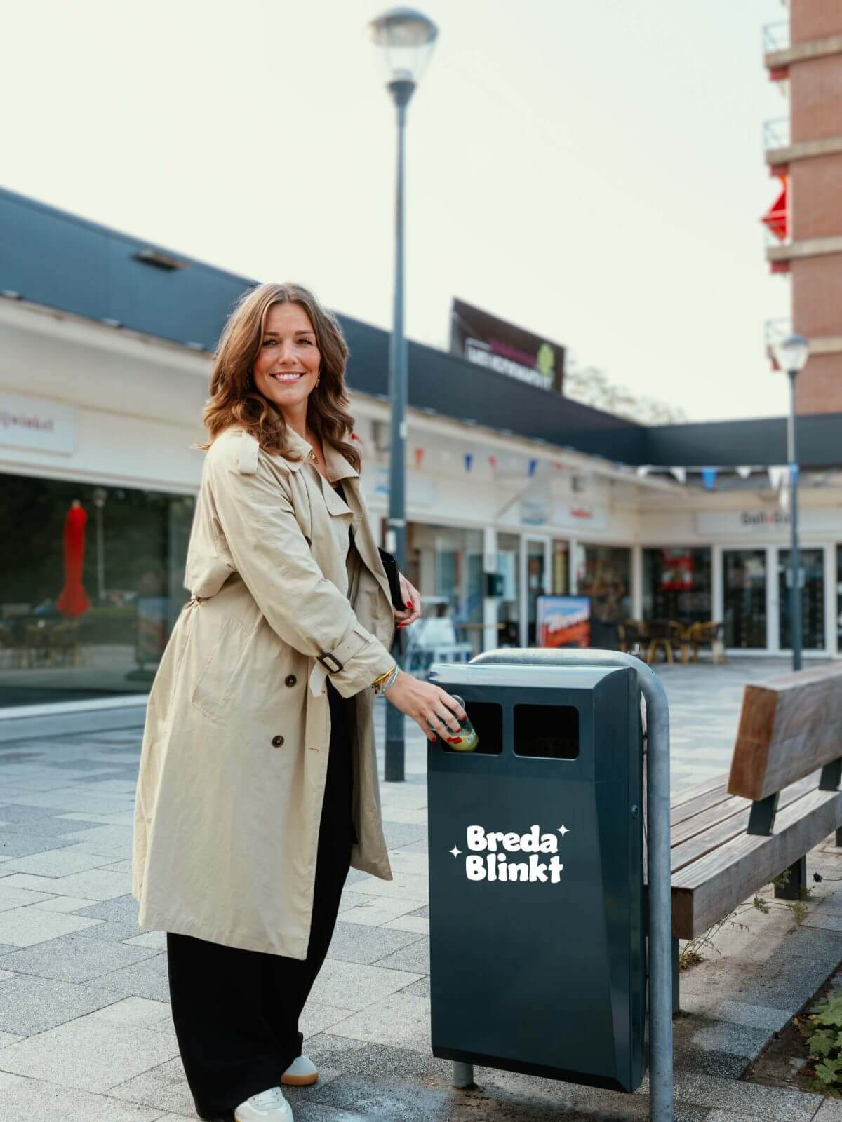 Lachende vrouw in beige jas gooit afval in een Breda Blikt afvalbak in een stadsstraat.