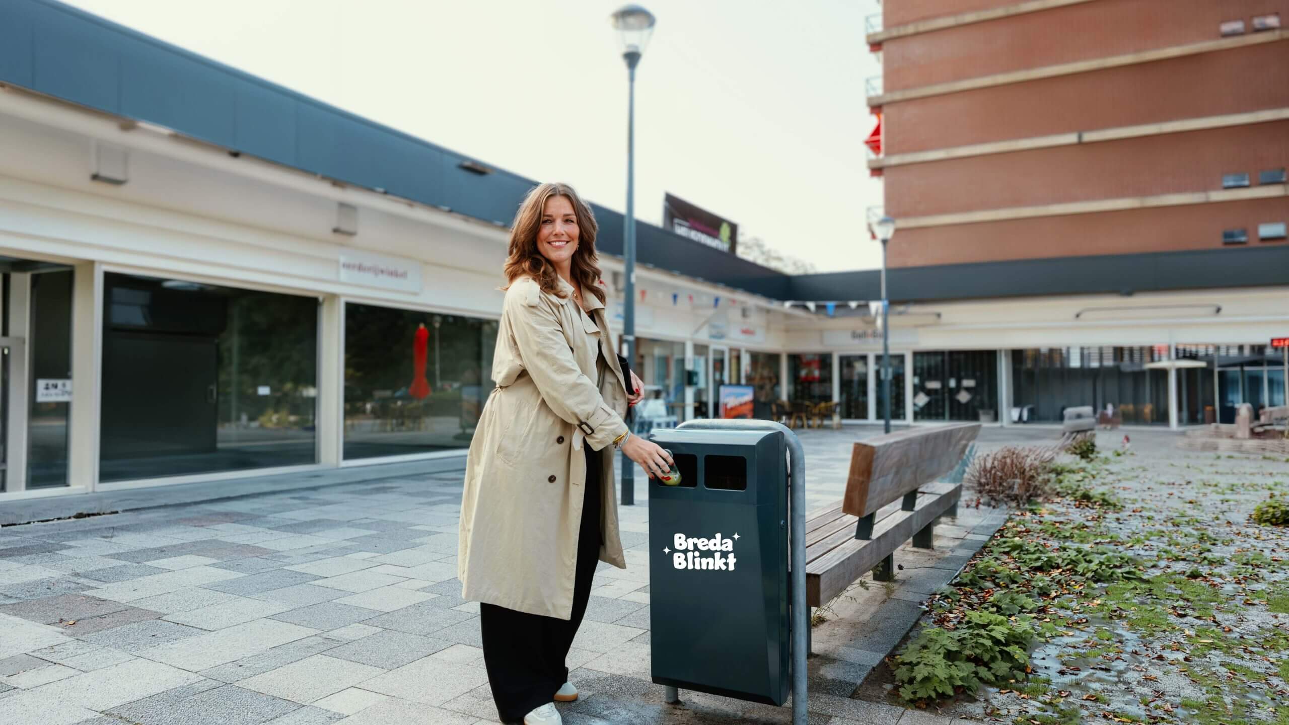 Lachende vrouw in beige jas gooit afval in een Breda Blikt afvalbak in een stadsstraat.