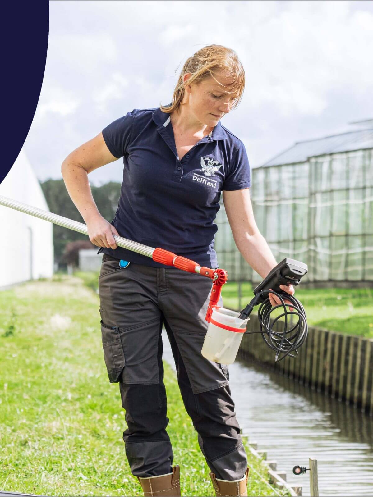 Vrouw in werkkleding bemonstert water uit een kanaal naast kassen op een zonnige dag.