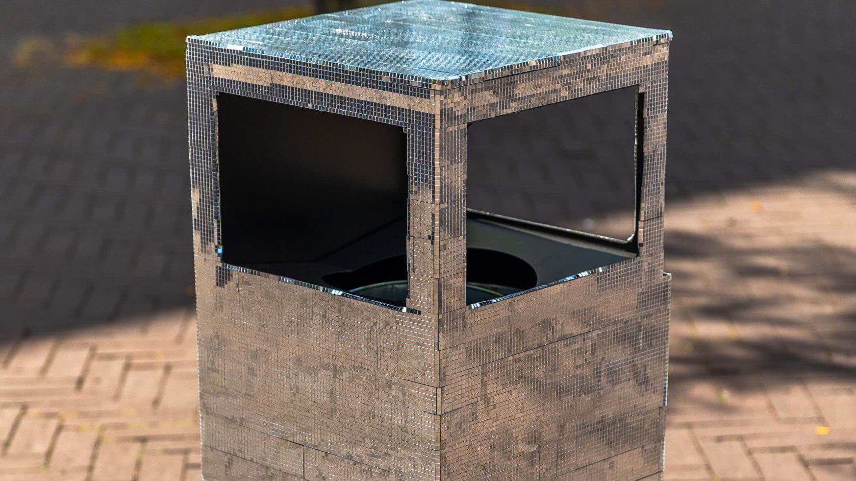Een zilveren metalen vuilnisbak met open zijkanten staat op een stenen stoep buiten.