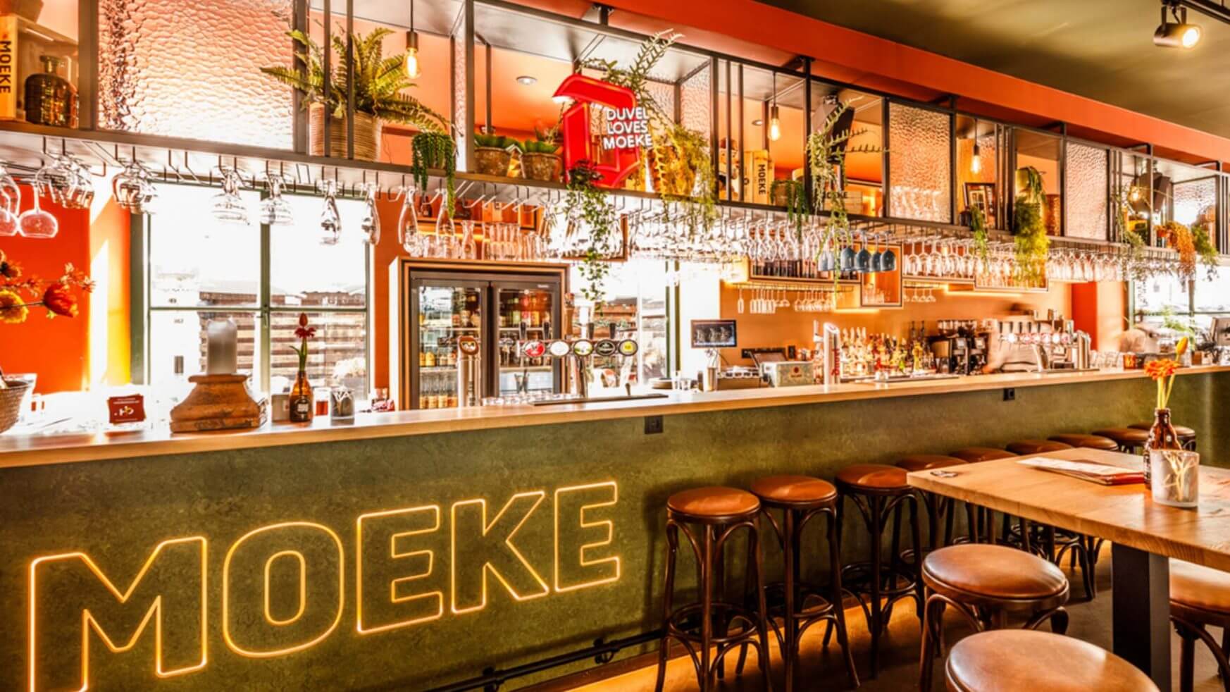 Strakke bar met "MOEKE" in neon, hoge krukken, hangende glazen en groen boven de toonbank.
