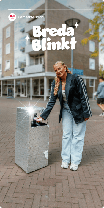 Lachende vrouw gebruikt een glimmende prullenbak in een stadsstraat, onder het bord "Breda Blinkt".