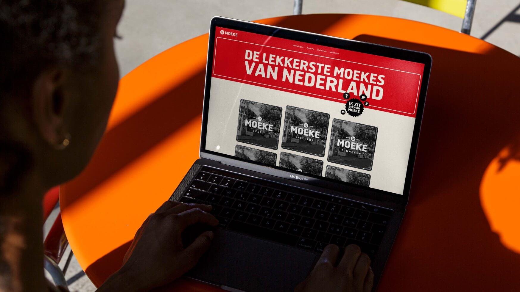 Persoon met een laptop aan een oranje tafel, bekijkt een website met Nederlandse tekst en afbeeldingen op het scherm.