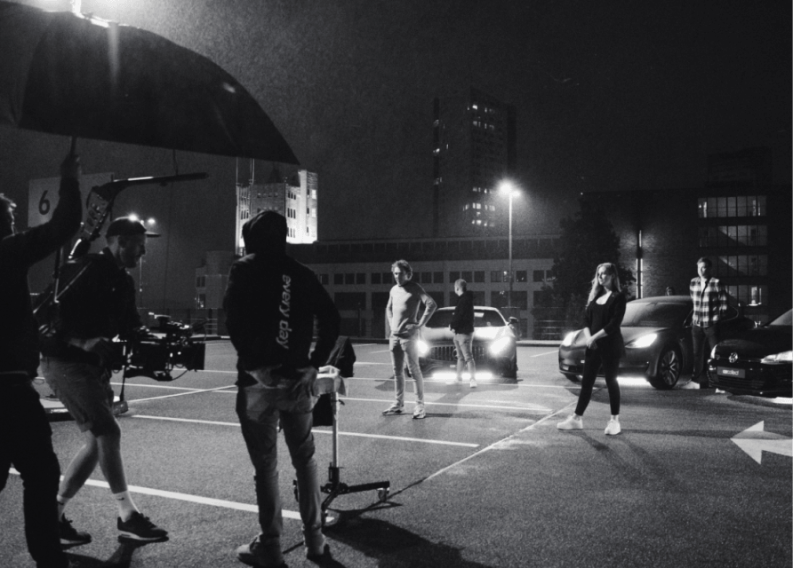 Filmploeg en acteurs filmen een nachtscène op een parkeerplaats in de stad, auto's en lichten zichtbaar, alles in zwart-wit.