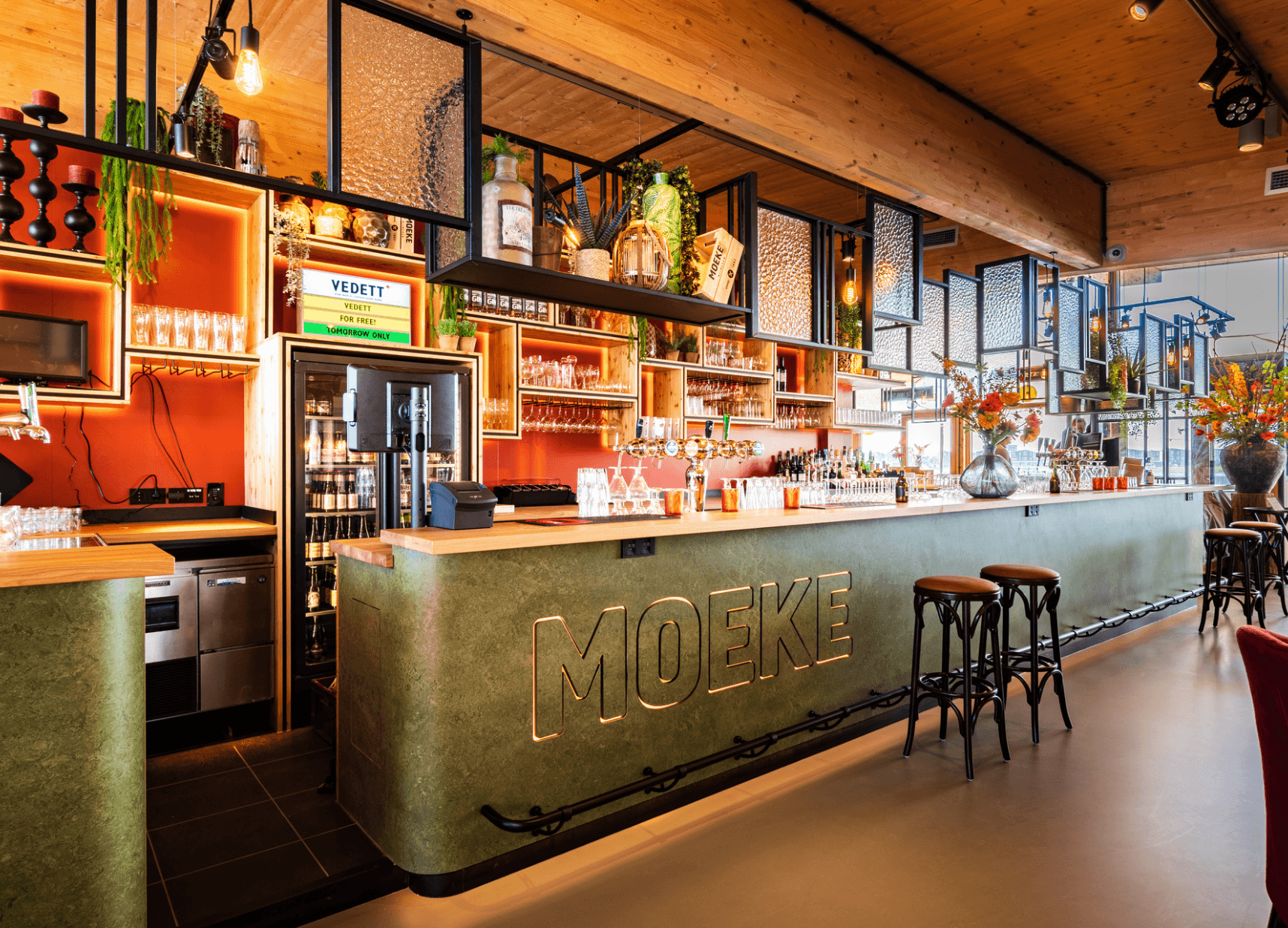 Chique bar met een groene MOEKE toonbank, planken met flessen en gezellige sfeerverlichting.