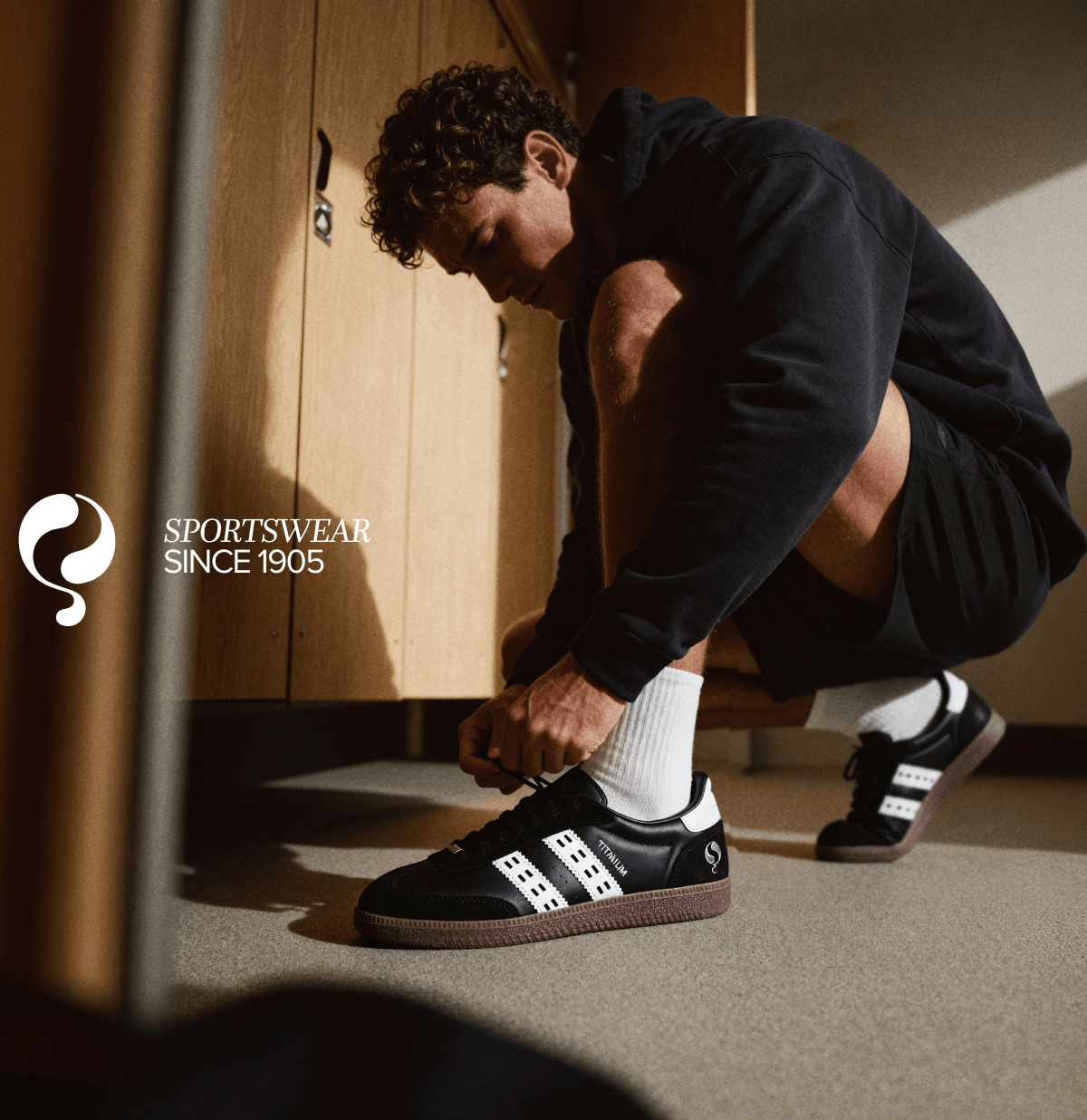 Atletische man knoopt zwarte gympen vast in een kleedkamer, met de tekst SPORTSWEAR SINCE 1905 en het merklogo zichtbaar.